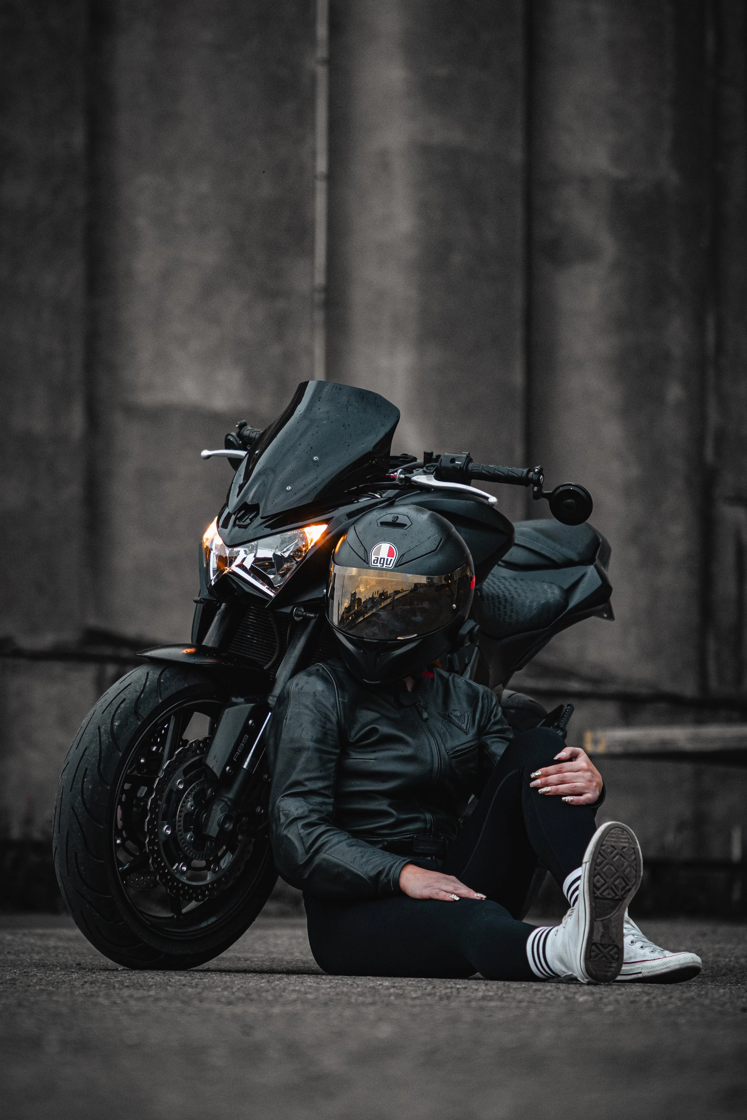 Person sitting on ground with a black motorcycle, wearing a black leather jacket, black pants, white sneakers, and a black motorcycle helmet with reflective visor