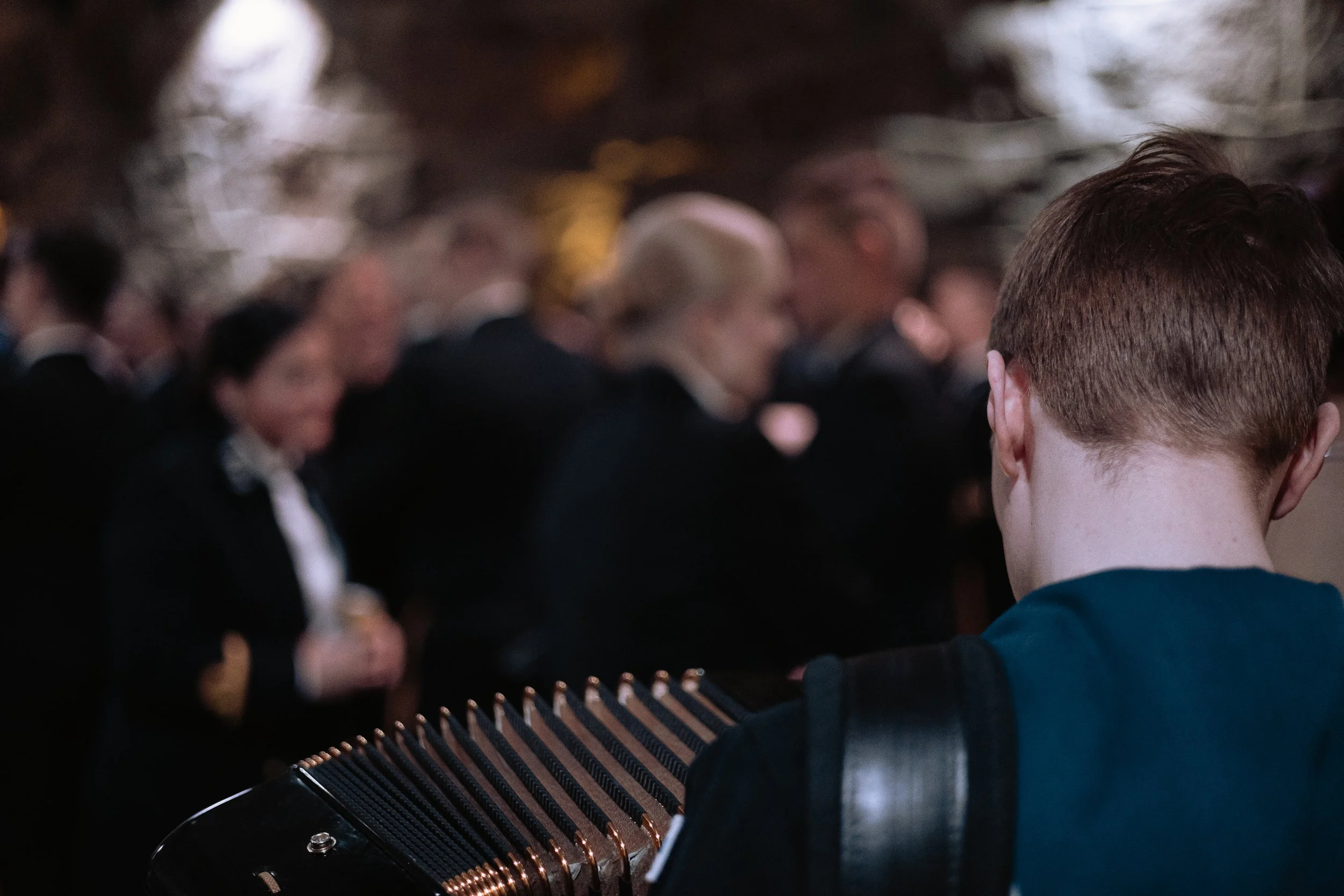 A young man playing the accordion at a formal event with blurred people in suits in the background.