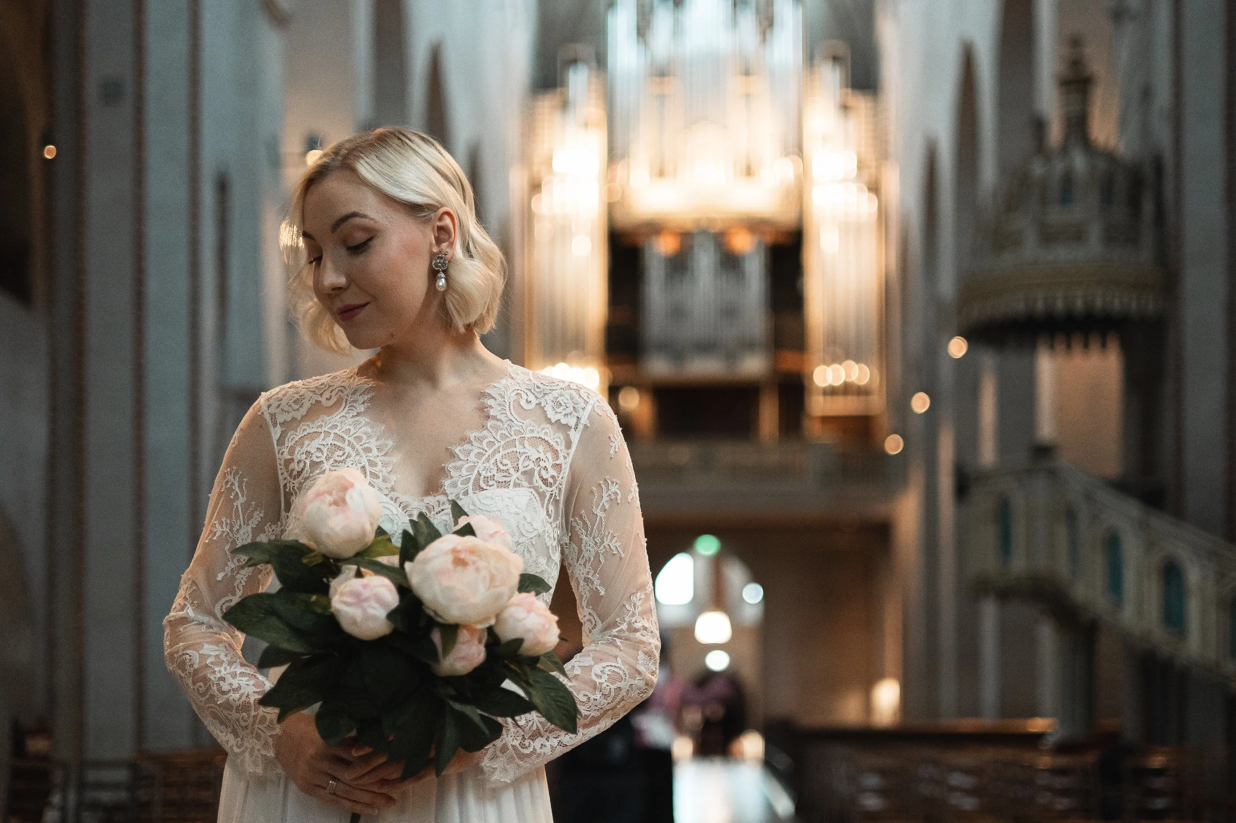 A bride in a lace wedding dress holding a bouquet of pink and white peonies inside a church with ornate architecture and organ in the background.