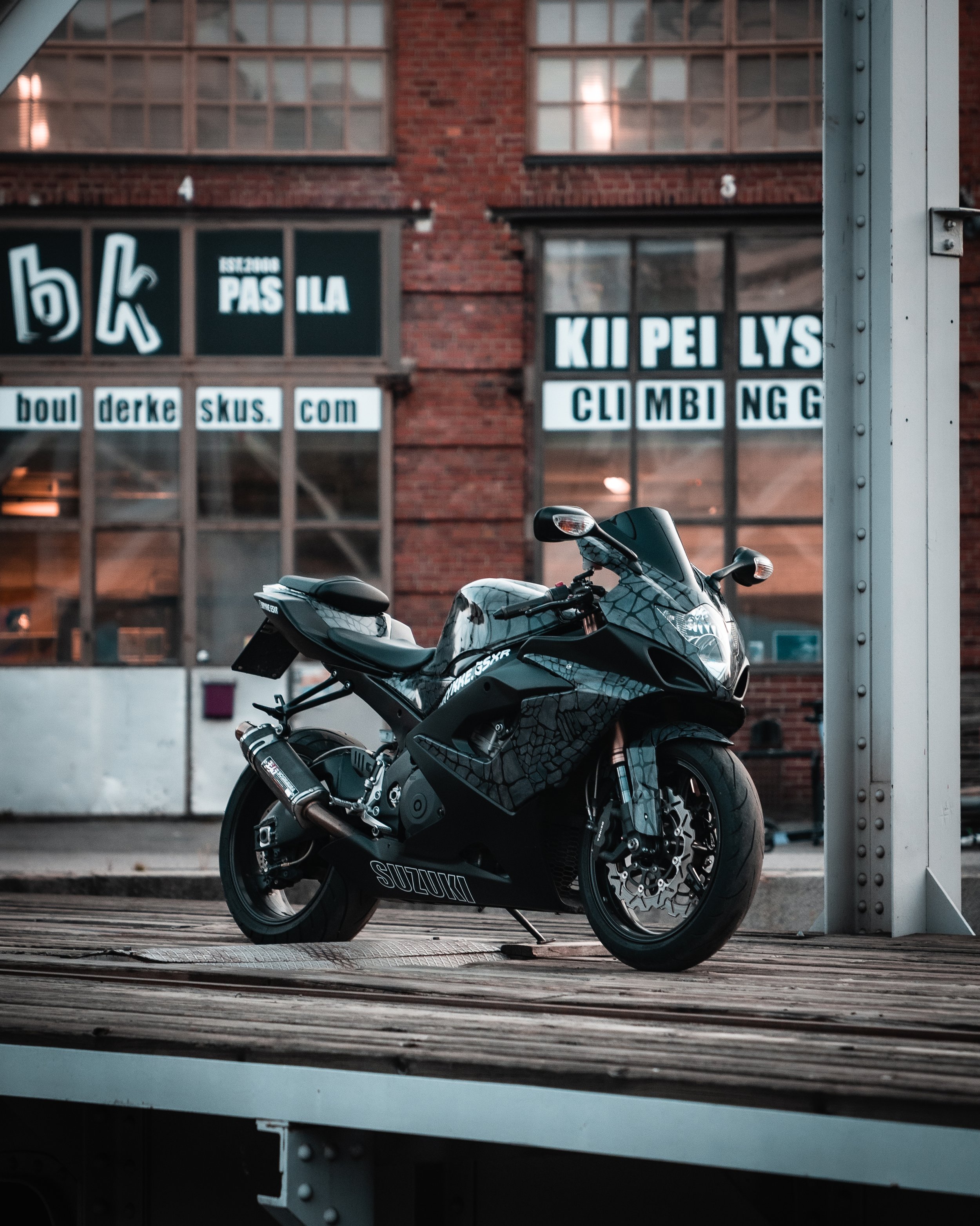 Black Suzuki motorcycle parked on a wooden platform in front of a brick building with large windows and white signs.