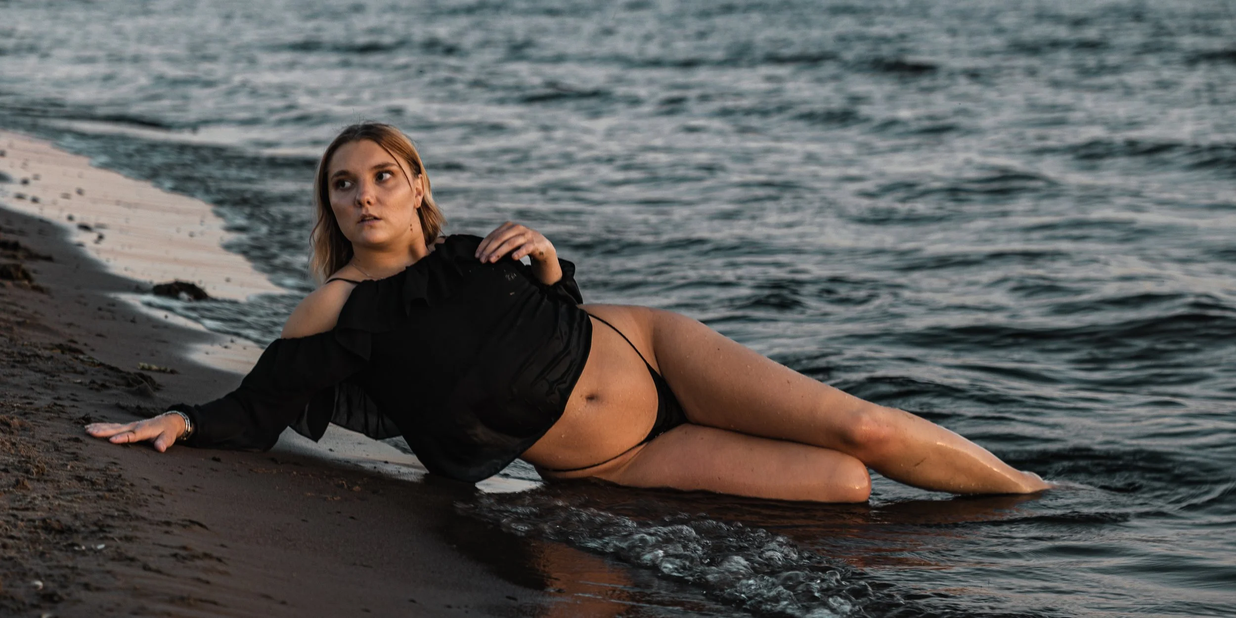 A woman in black clothing lying on the shoreline with water reaching her waist, looking to the side with an expression of surprise or concern.