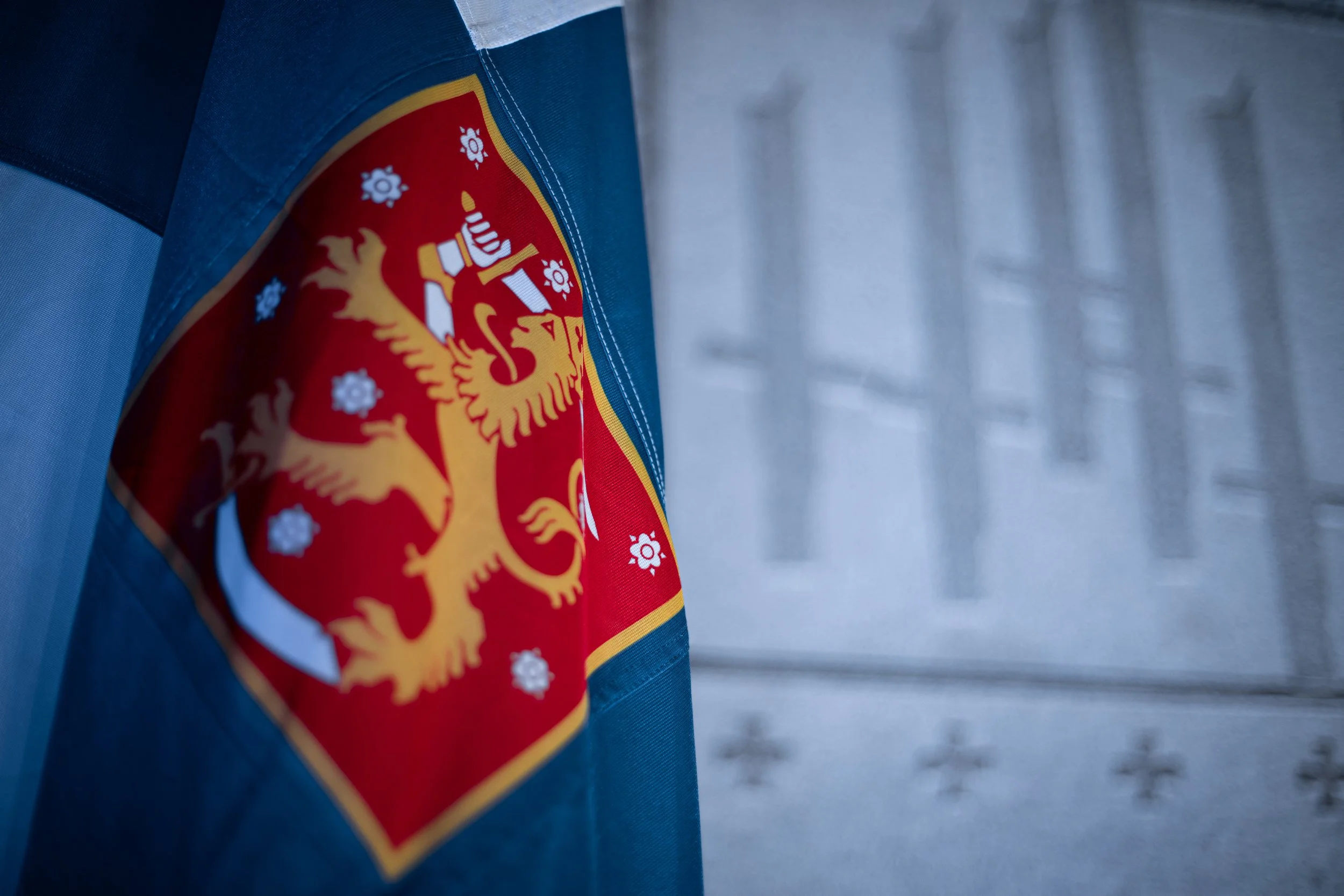 Close-up of a flag with a coat of arms featuring a crowned lion, with a building featuring columns in the background.