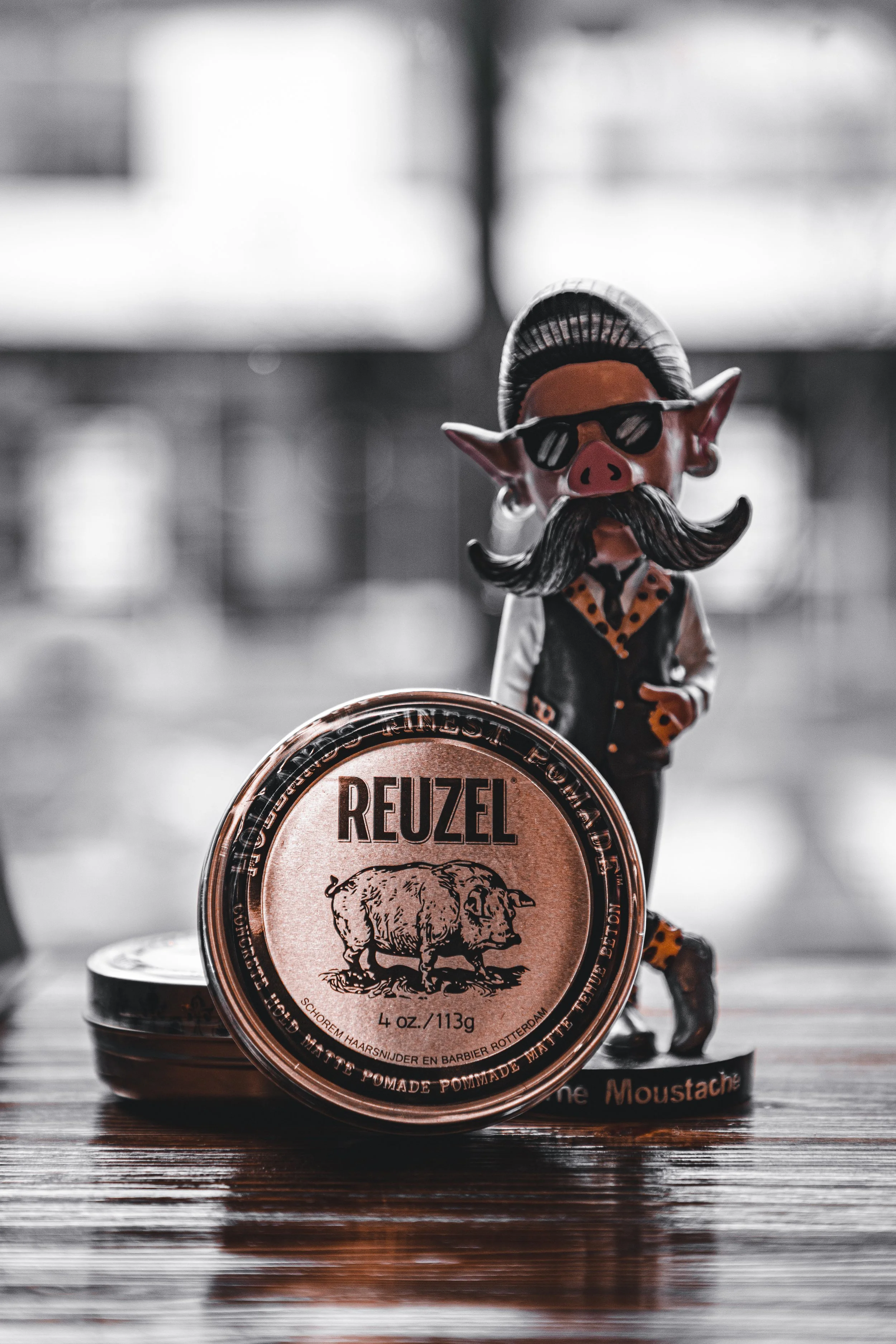 A toy figure of a pig with a mustache, sunglasses, and a hairstyle, standing next to a tin of Reuzel pomade on a wooden surface, with a blurred background.