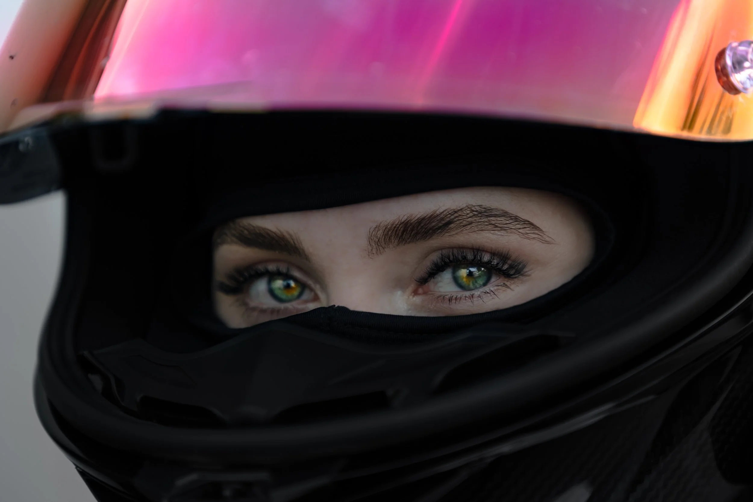 Close-up of a woman with blue-green eyes wearing a black motorcycle helmet with a reflective visor in a pink-orange gradient.
