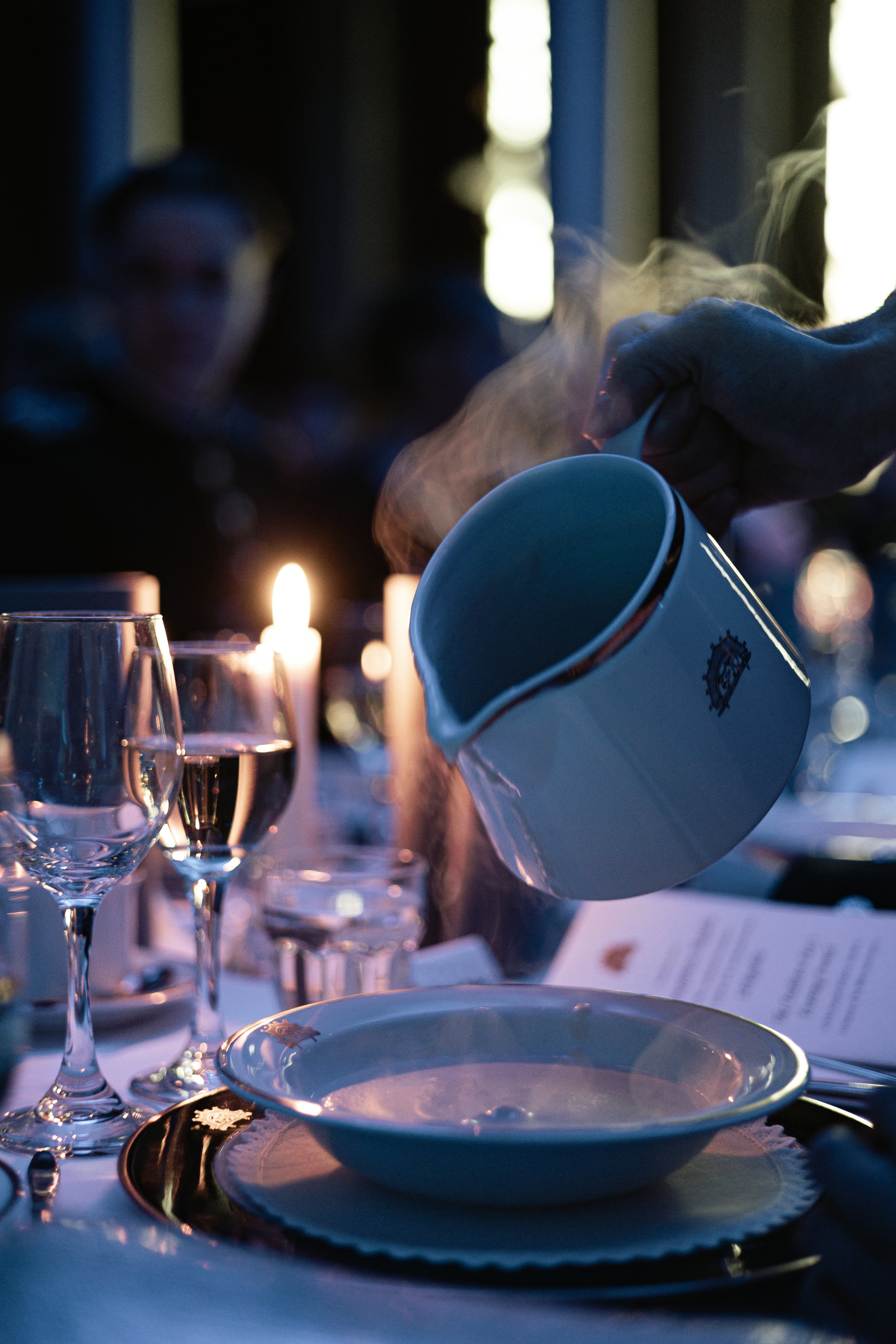 A person pouring hot coffee from a white mug into a bowl on a formal dining table set with wine glasses, candles, and plates during evening or early night.