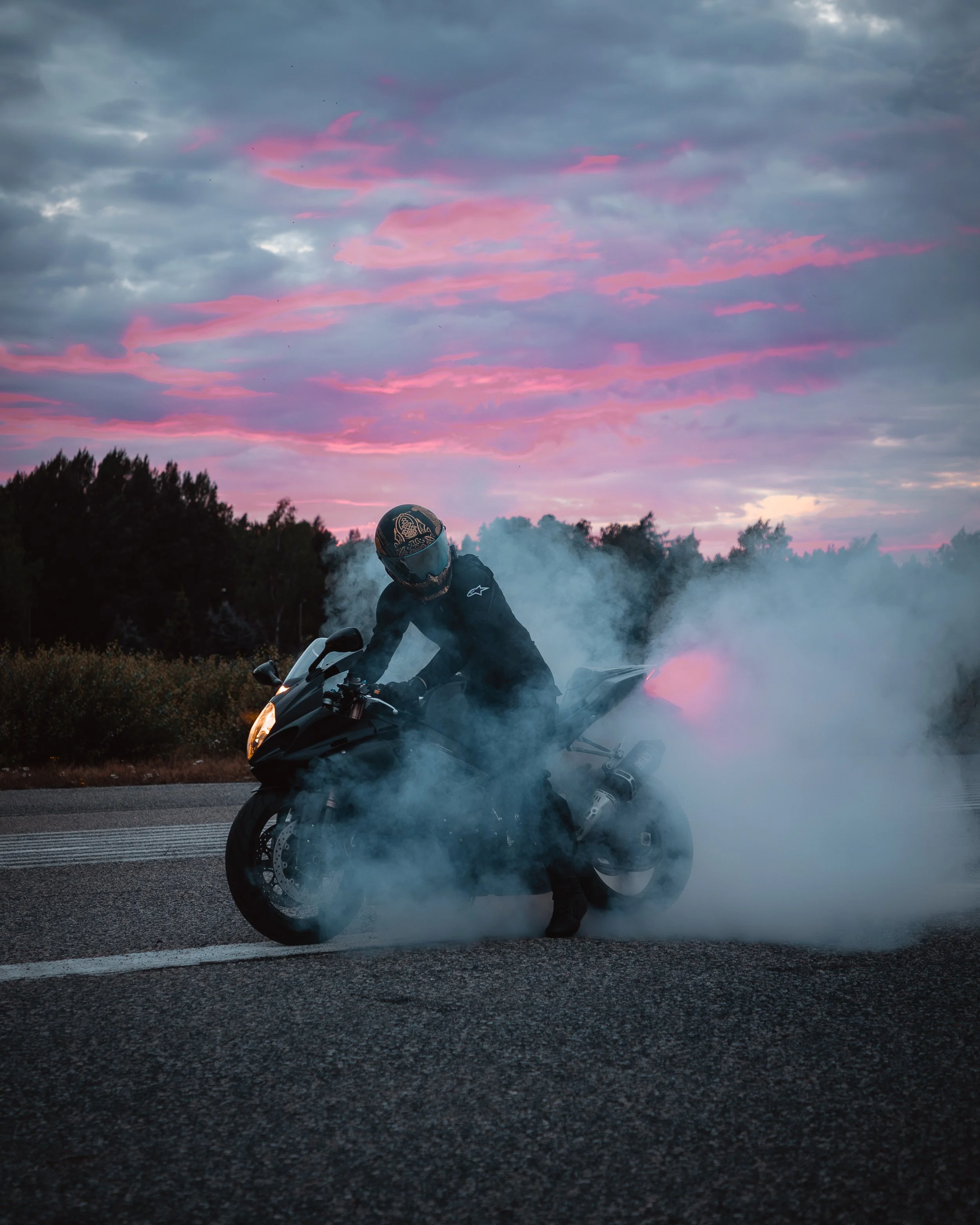 Motorcycle rider in dark gear doing a burnout on the road during sunset, with colorful clouds and trees in the background.