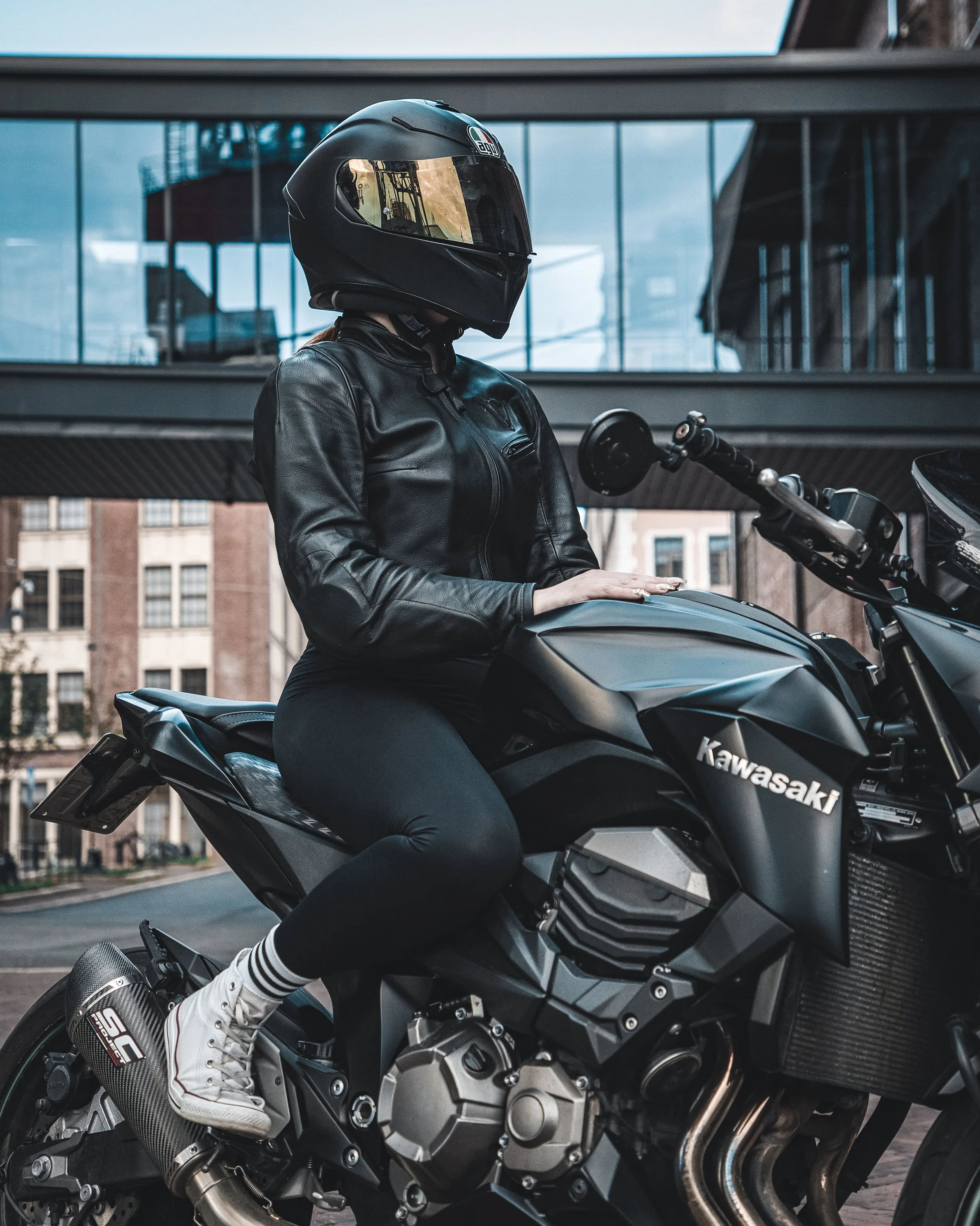 A person wearing a black motorcycle helmet, black leather jacket, black pants, and white sneakers sitting on a black Kawasaki motorcycle.