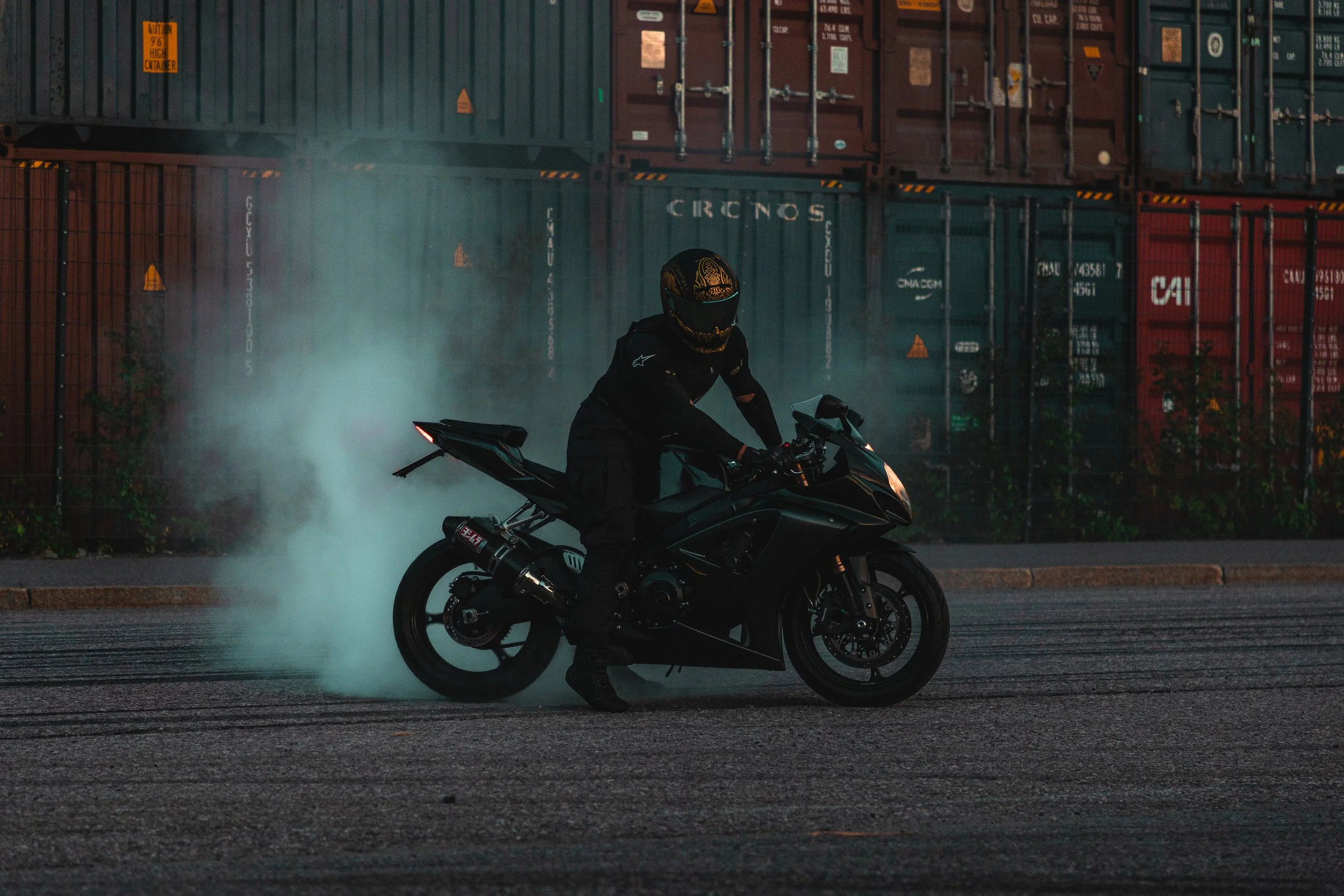 A motorcyclist wearing black gear and a gold helmet doing a burnout on a black sportbike, emitting smoke from the rear tire, on an empty street with trucks and shipping containers in the background.