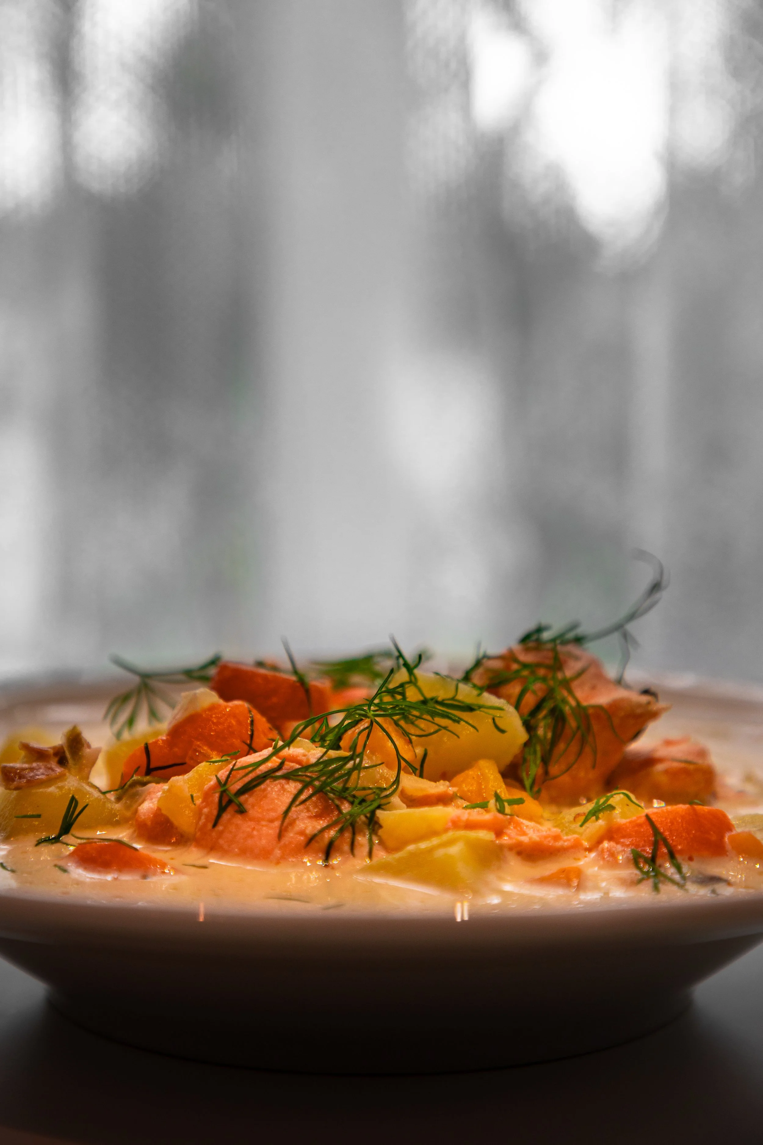 A bowl of creamy fish chowder garnished with fresh dill, containing chunks of salmon, potatoes, and carrots, set against a blurred window background.