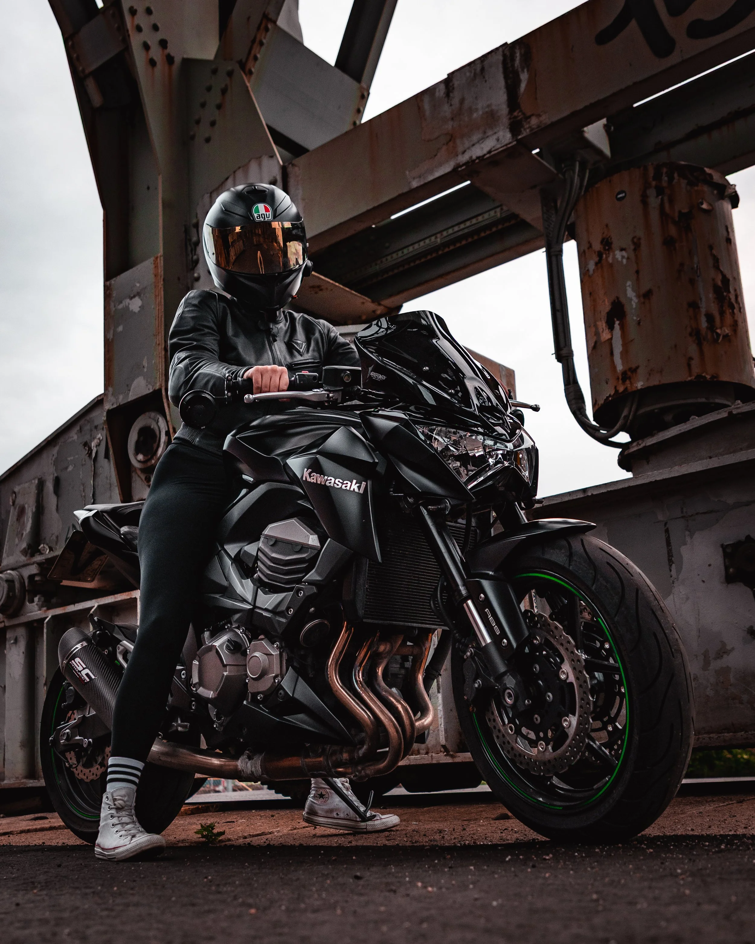 A person dressed in black motorcycle gear, including a helmet and jacket, sitting on a sleek black Kawasaki motorcycle under a rusted steel bridge structure.