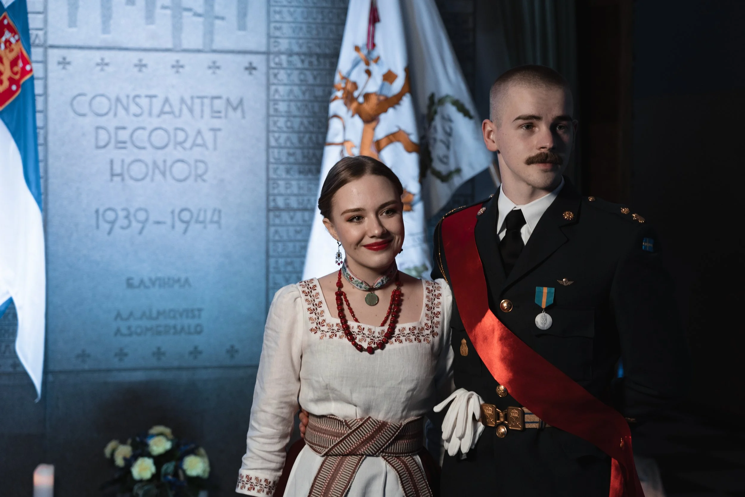 A woman and a man in military uniform standing together at a memorial, with flags and a plaque commemorating World War II veterans in the background.