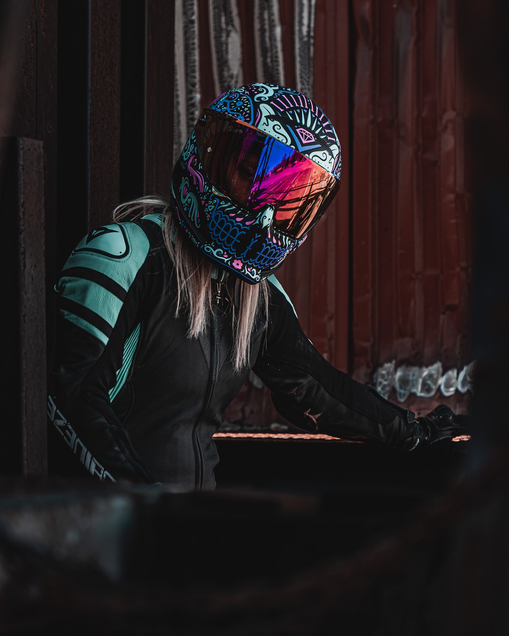 Person wearing a colorful skull pattern helmet and motorcycle gear leaning against a dark wall with a rusty red background.