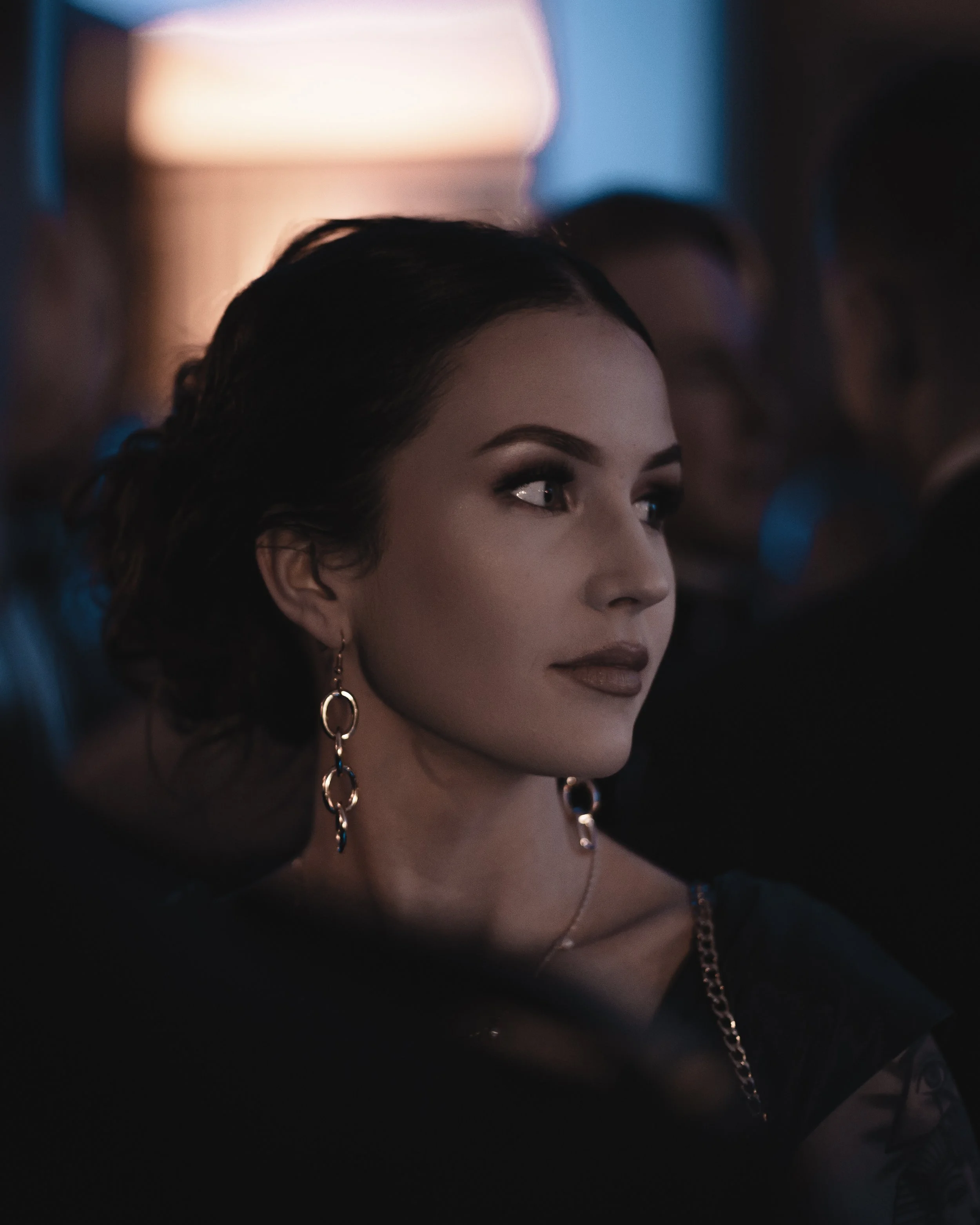 A woman with short dark hair and earrings looking to her left in a dimly lit indoor setting with people in the background.