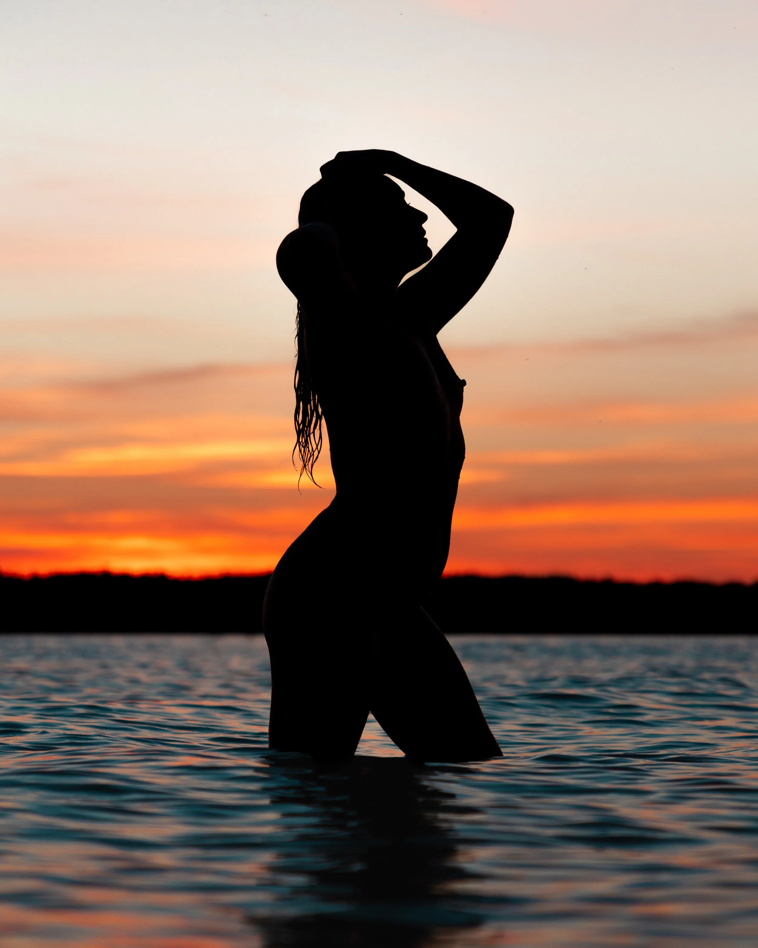 Silhouette of a woman standing in water during sunset with her hand on her head.