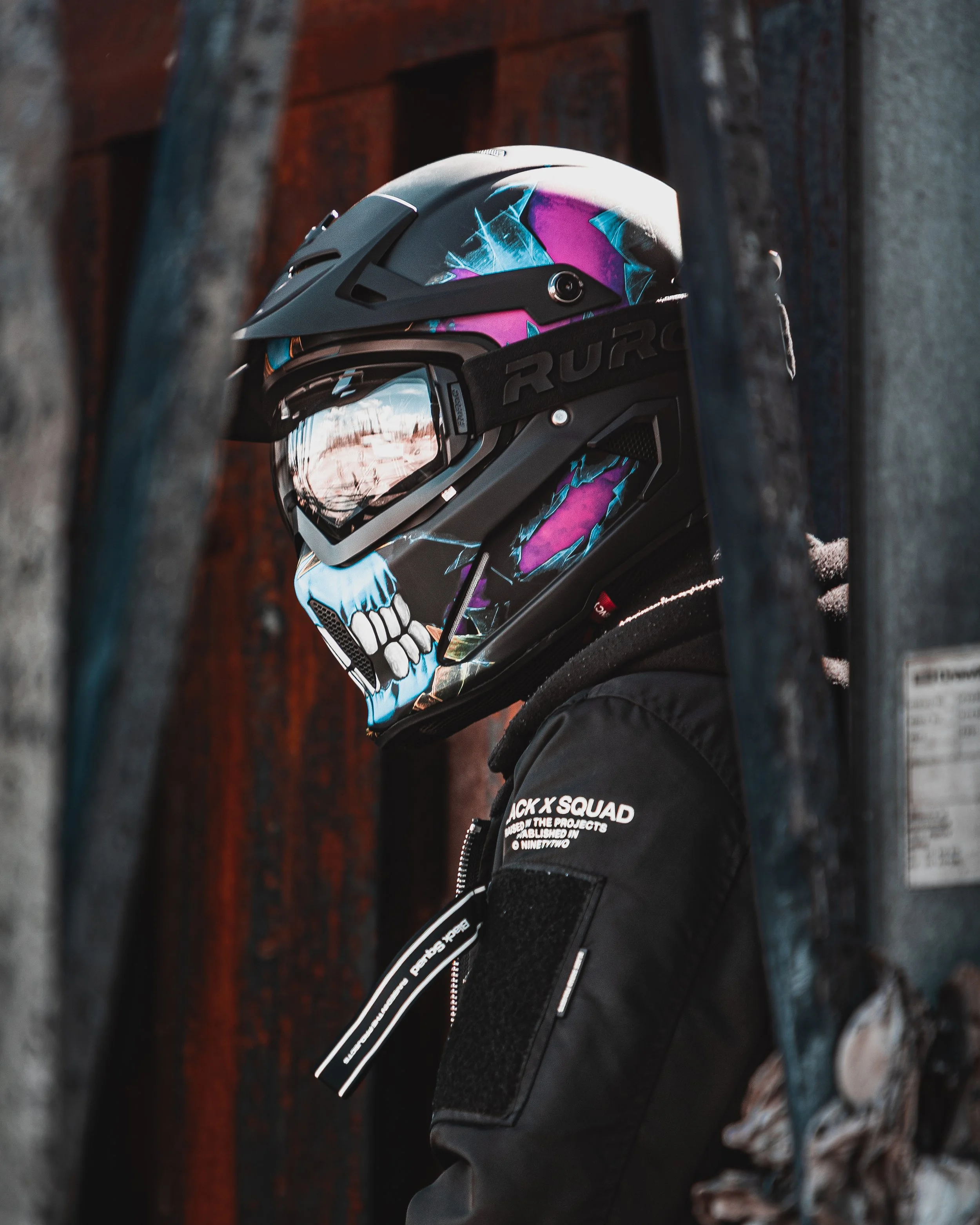 Person wearing a black jacket and a colorful skull-themed motorcycle helmet, standing near a chain link fence with a wooden wall in the background.