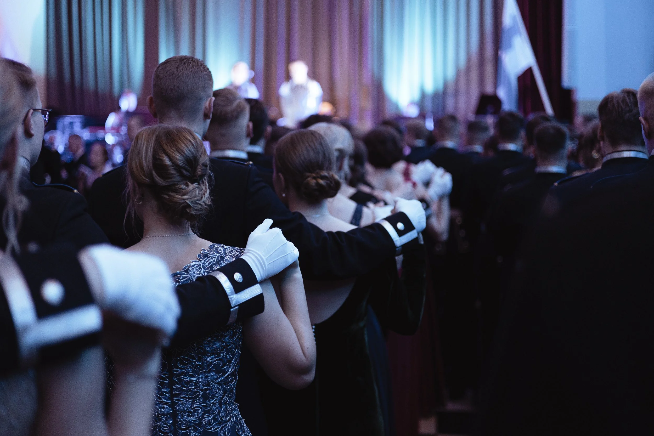A formal ballroom dance event with couples in elegant attire, including women in dresses and men in tuxedos, dancing in a line with their arms around each other's shoulders, in front of a stage with a speaker and a flag.
