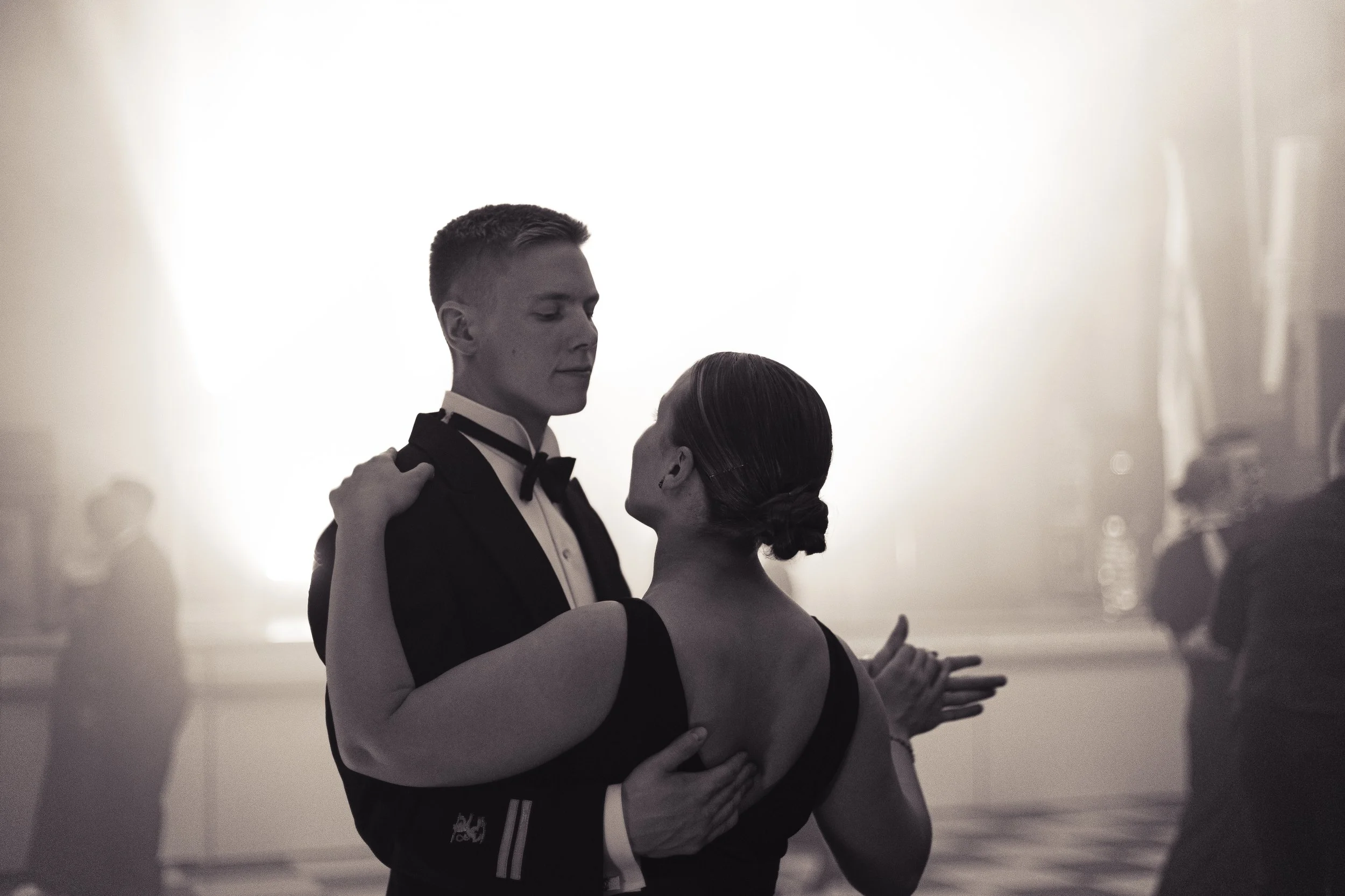 A young man and woman dancing closely at a formal event, with other couples dancing in the background, in black and white.