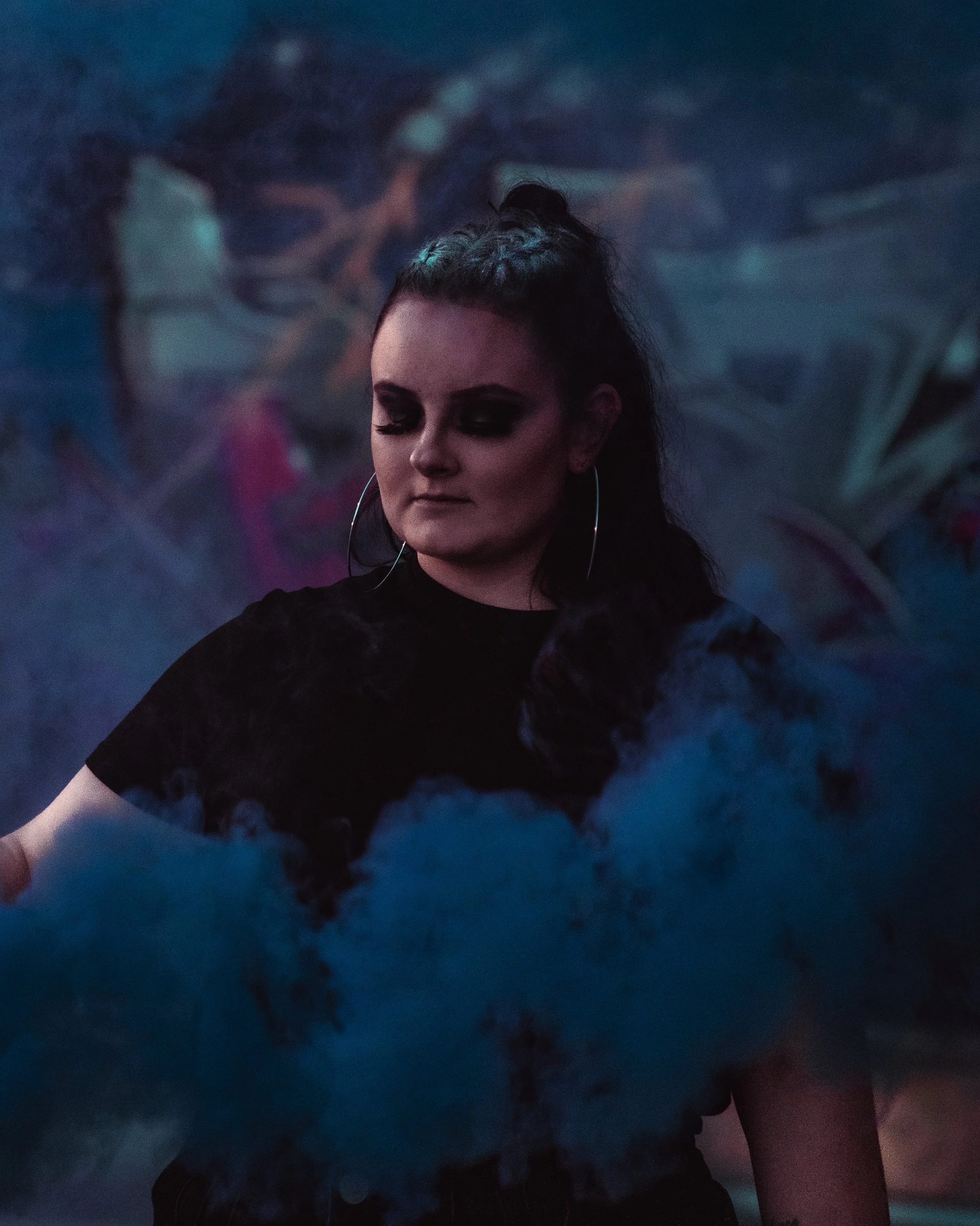 A young woman with dark hair and eye makeup stands amid blue smoke, wearing a black top and large hoop earrings in a dimly lit environment with graffiti background.