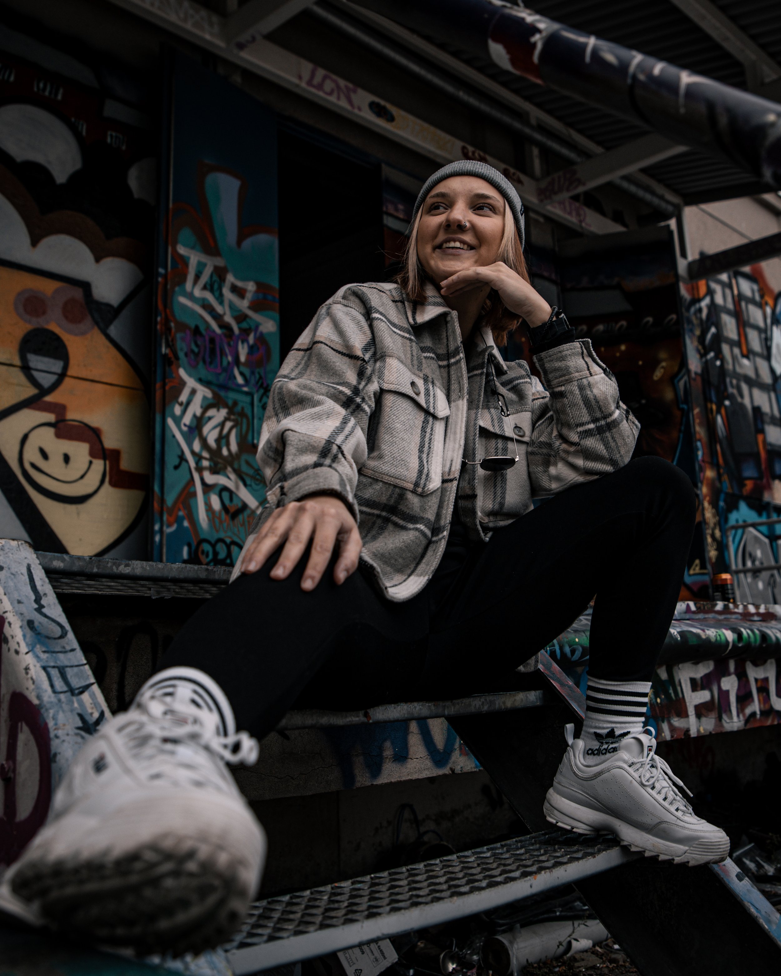 A young woman sitting on stairs at an outdoor skate park, surrounded by graffiti art, wearing a gray plaid jacket, black pants, white Adidas sneakers, and a gray beanie, smiling and looking to the side.