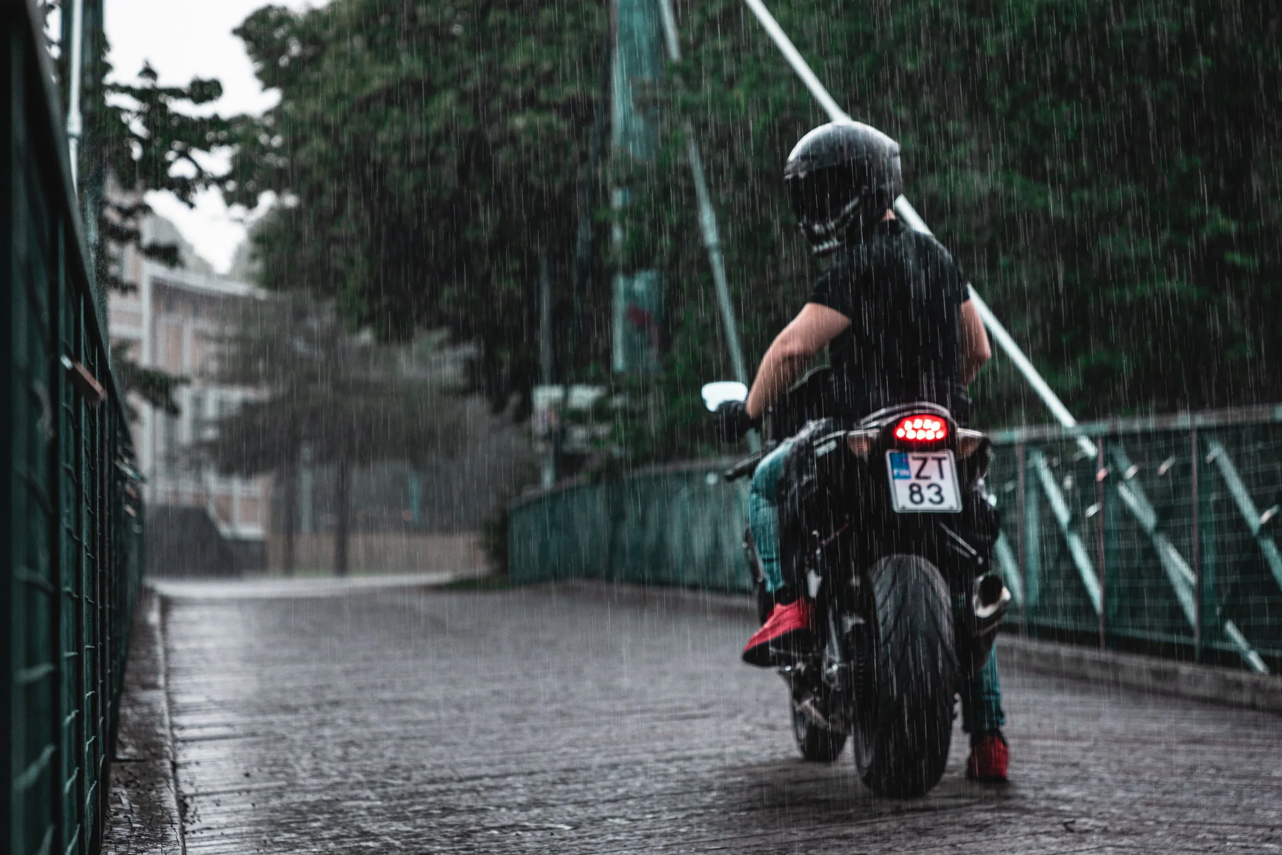 Motorcyclist riding a motorcycle in the rain on a paved street with trees and fences on the sides.