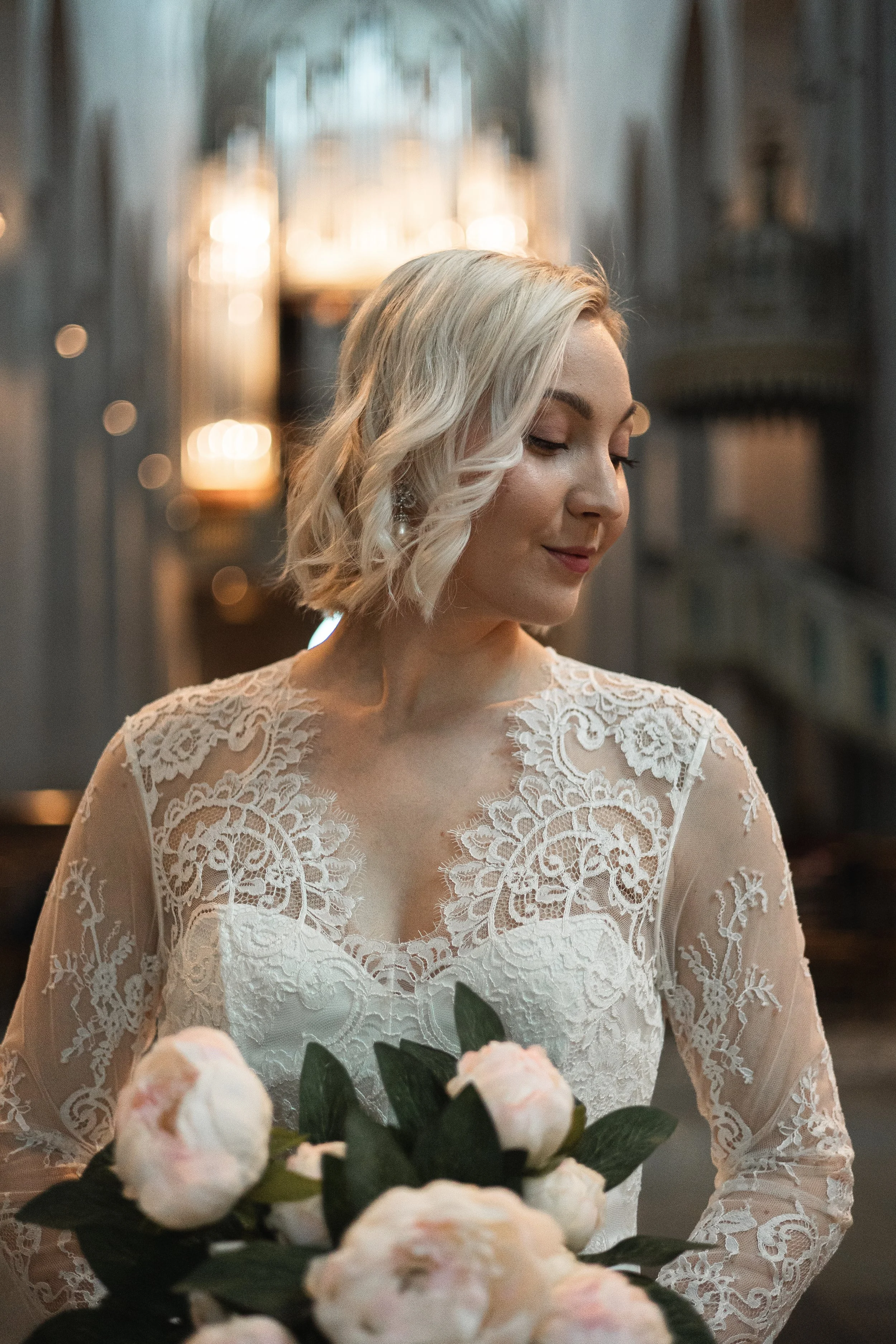 A woman in a lace wedding dress holding a bouquet of pink and white peonies, standing inside a church with soft lighting.