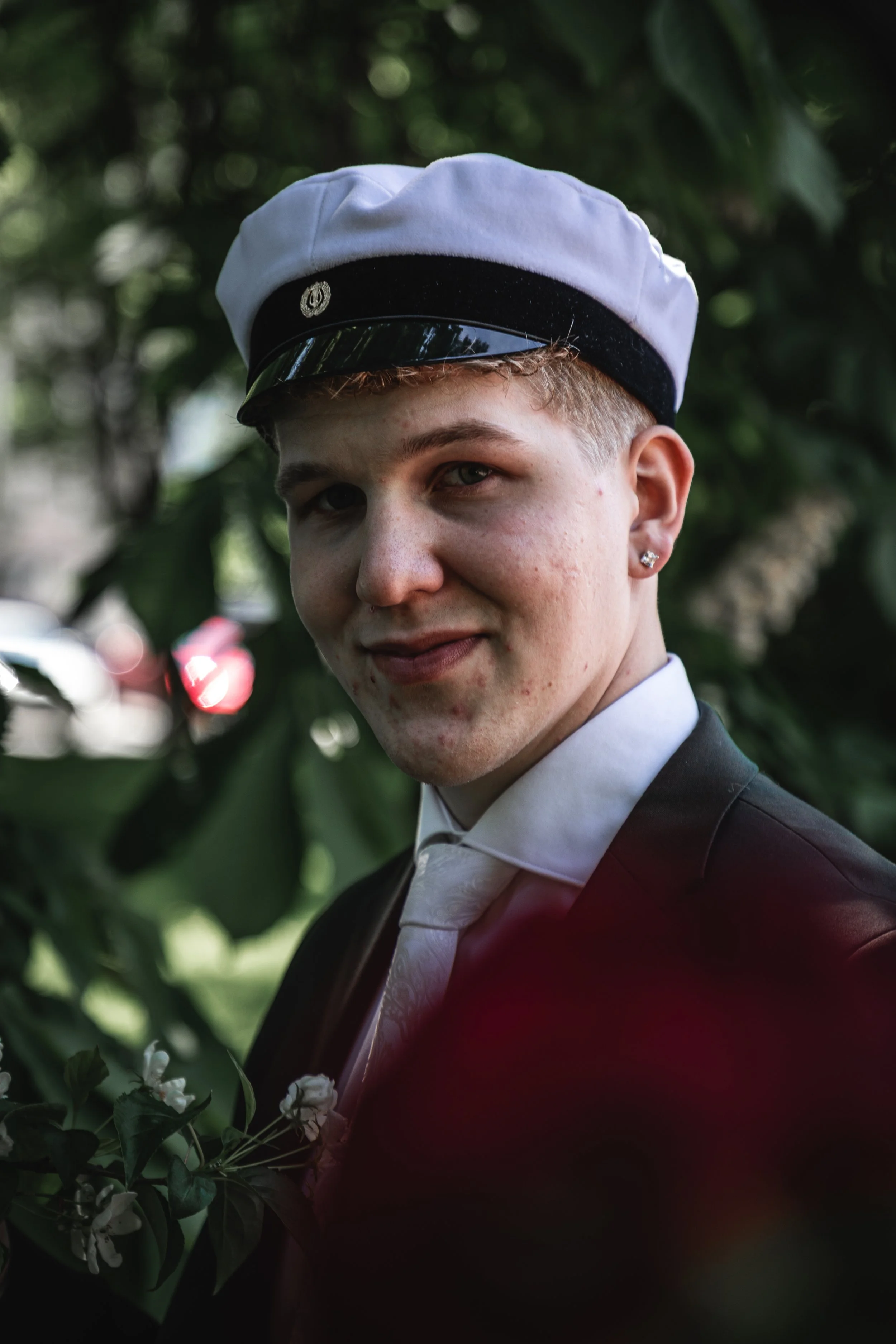 A young person with short, light hair wearing a white and black hat, white shirt, black suit, and a tie, standing outdoors with green foliage in the background.