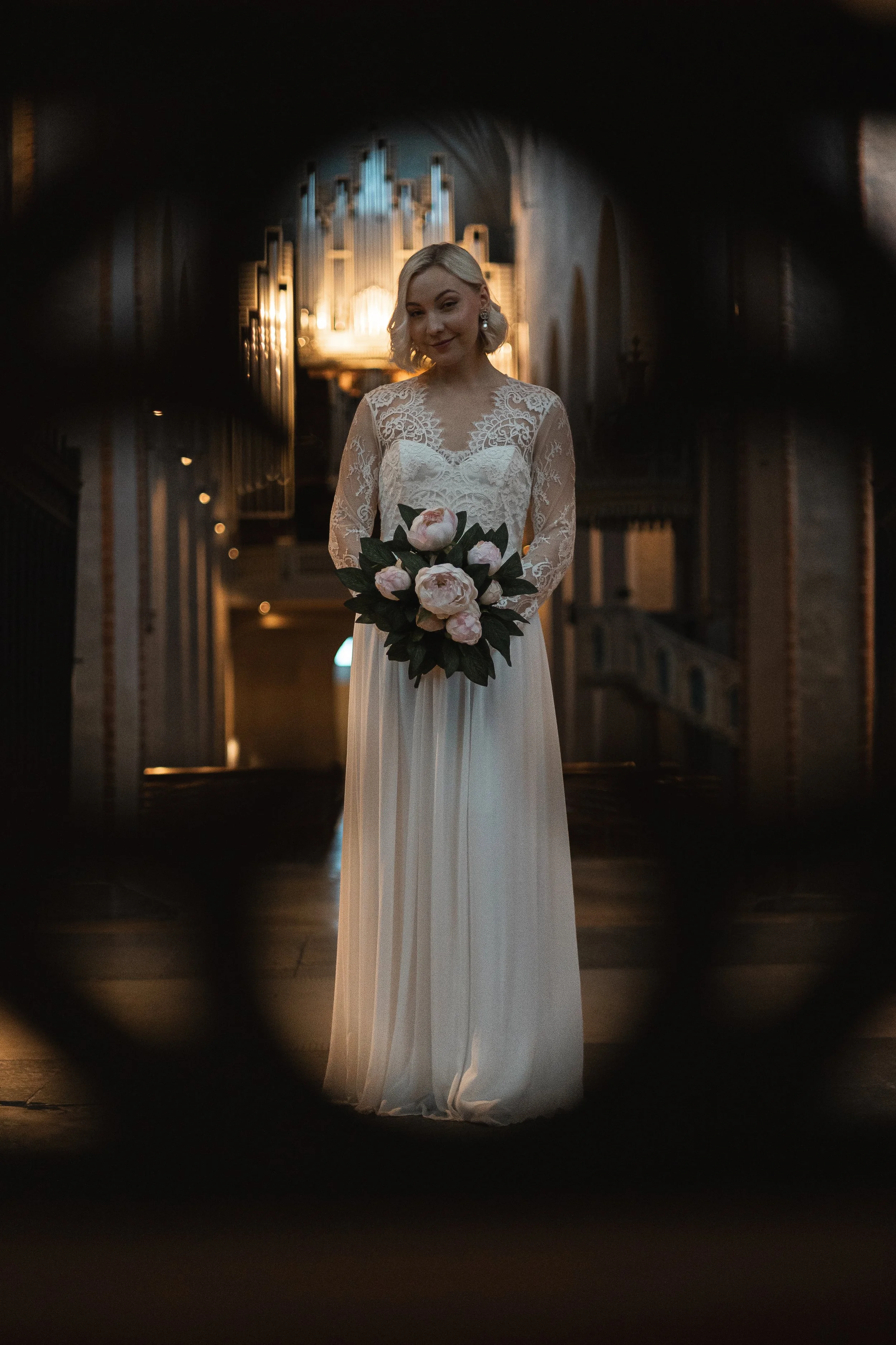 A bride in a lace wedding dress holding a bouquet of pink and white peonies, standing inside a church with ornate architecture and warm lighting.