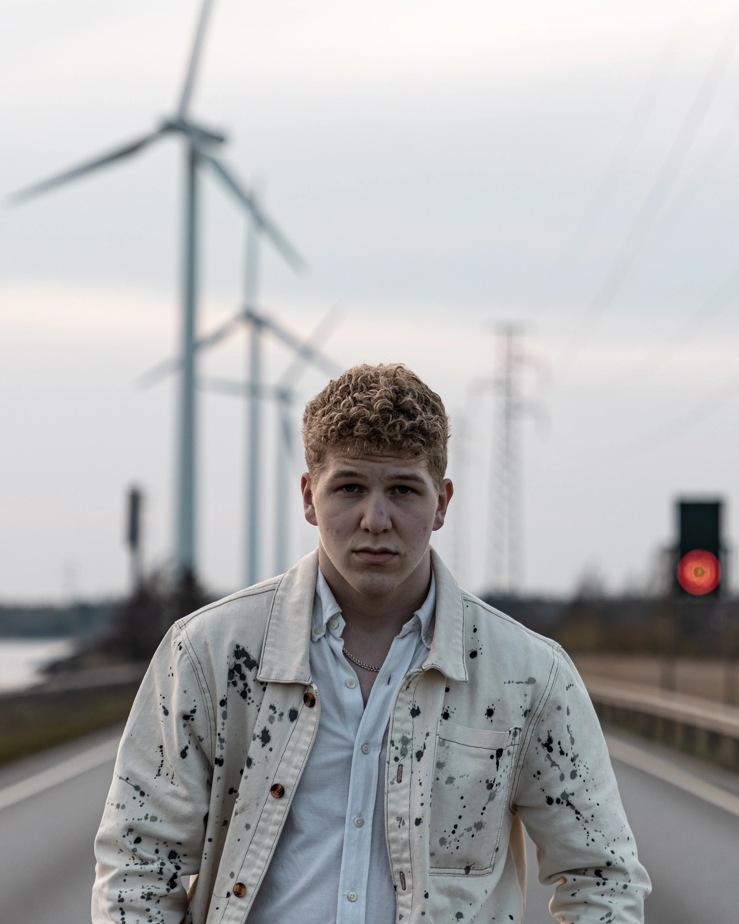 A young man with curly hair and a serious expression standing on an empty road with wind turbines in the background, wearing a paint-splattered beige jacket over a white shirt.