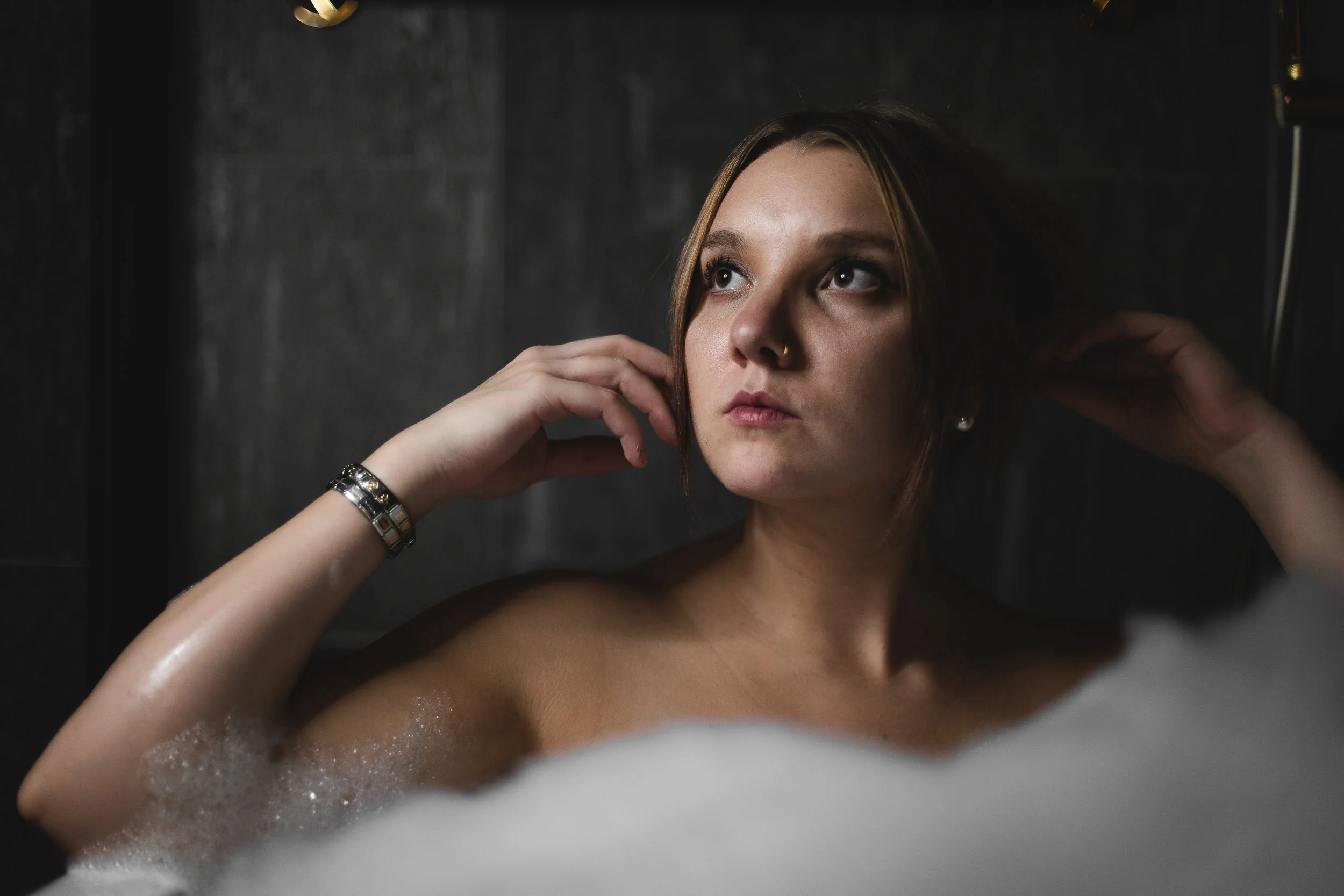 A young woman with light skin and shoulder-length brown hair relaxing in a bubble bath, adjusting her hair with her hands, looking contemplative. She is wearing a bracelet and pearl earrings, with a dark tiled wall in the background.