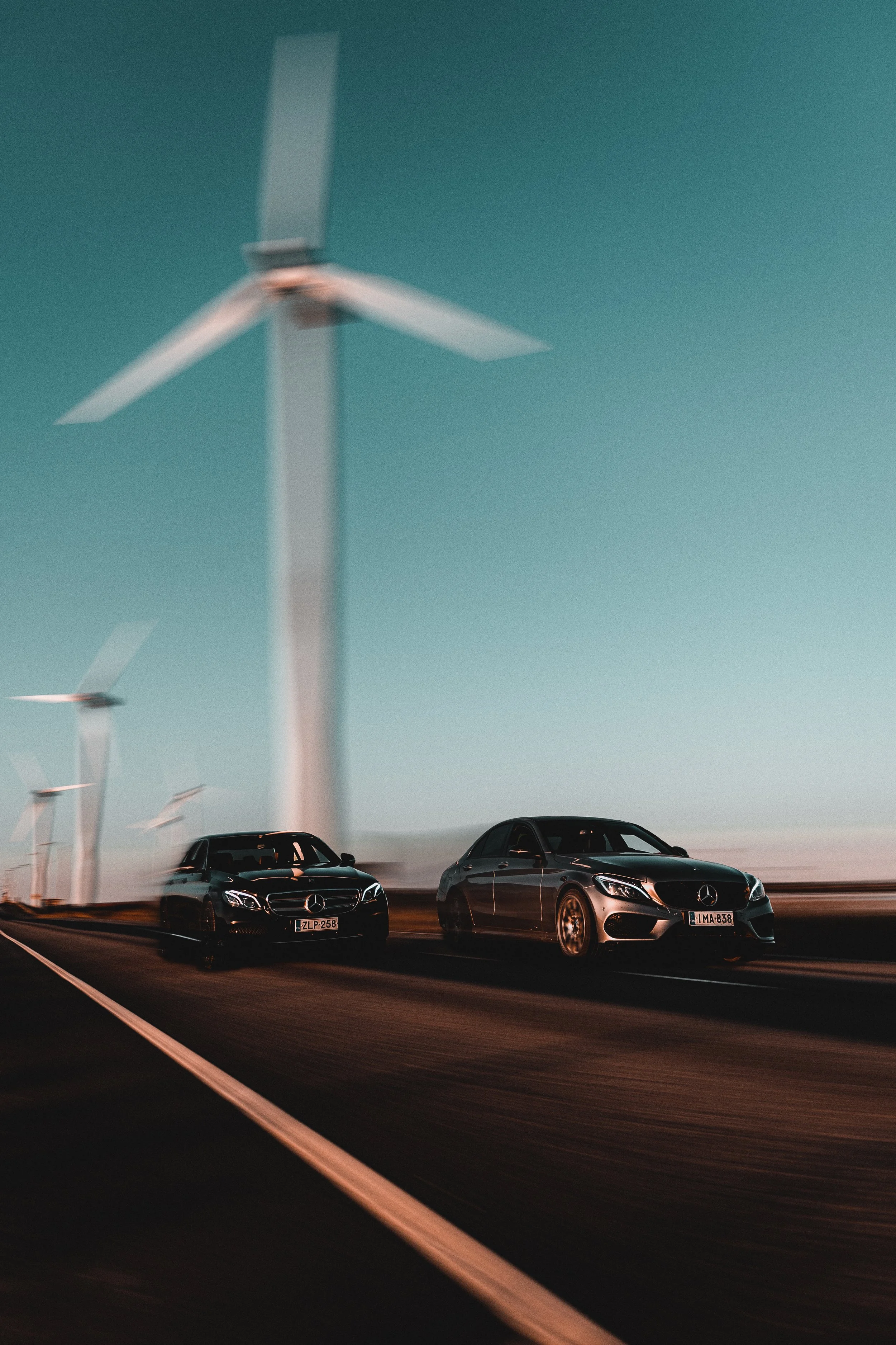 Two black Mercedes-Benz sedans driving on a road with wind turbines in the background during dusk or dawn.