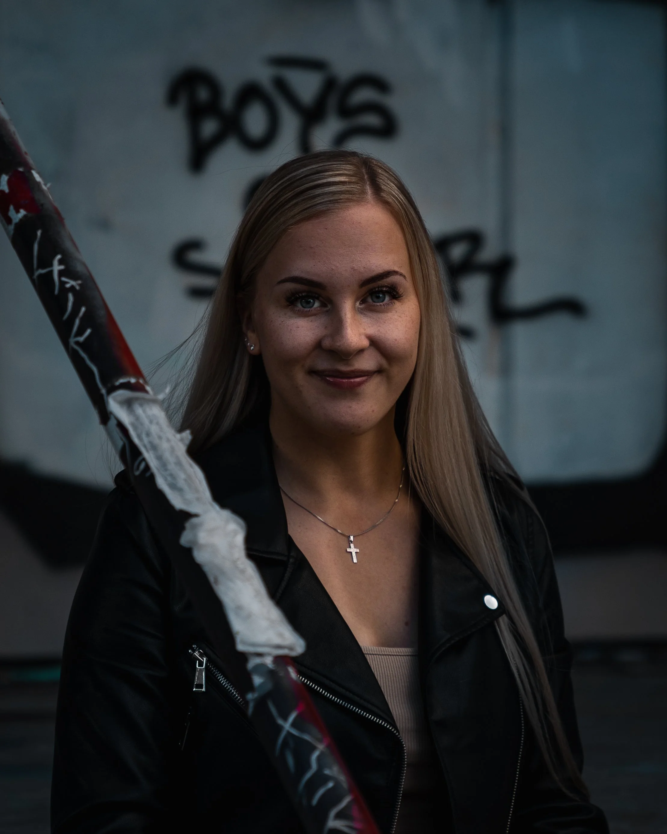 A woman with long blonde hair, wearing a black leather jacket and a cross necklace, standing in front of a wall with graffiti that reads 'BOYS'.