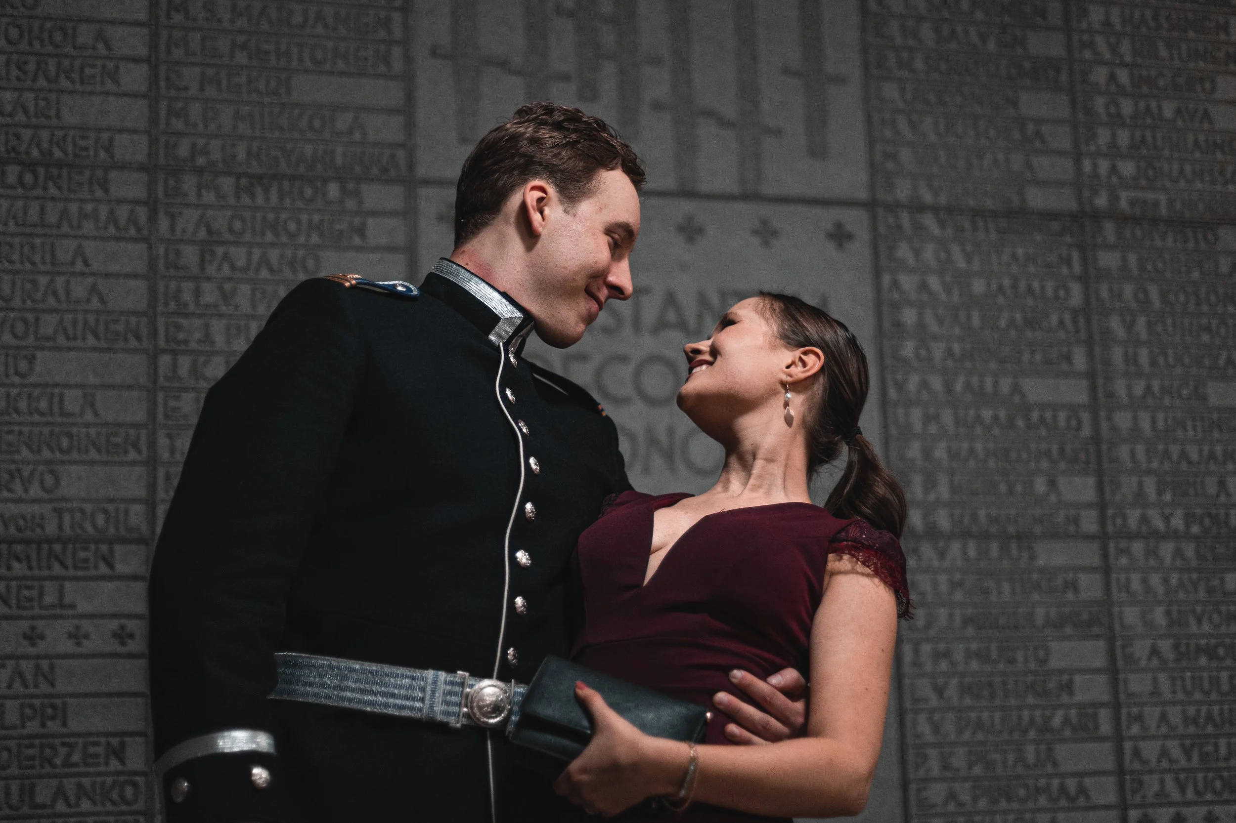 A couple dancing closely together, smiling, in front of a wall with engraved names.