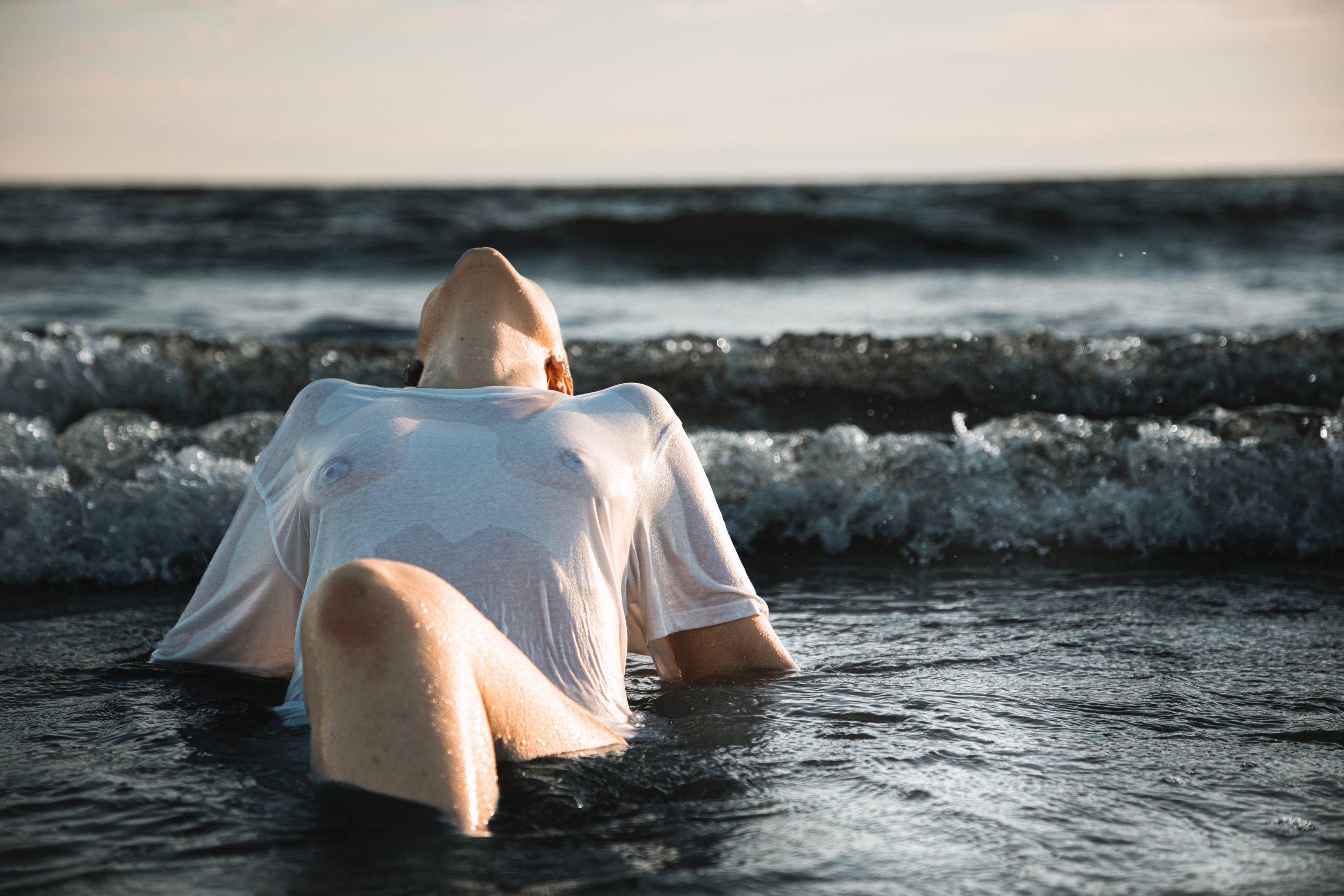 Person lying on their back in the ocean surf with head and upper body resting on the water, wearing a white shirt, in sunset lighting.