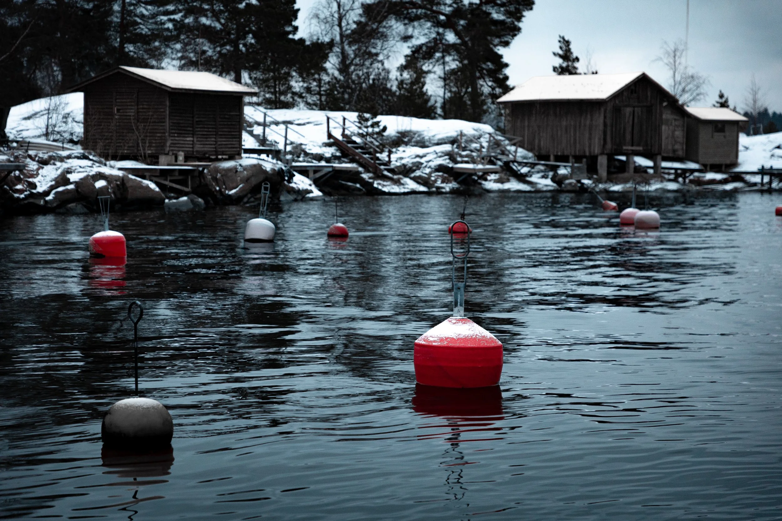 A winter lake scene with floating buoys tied to the water, snow-covered rocks, small wooden cabins, and trees in the background.