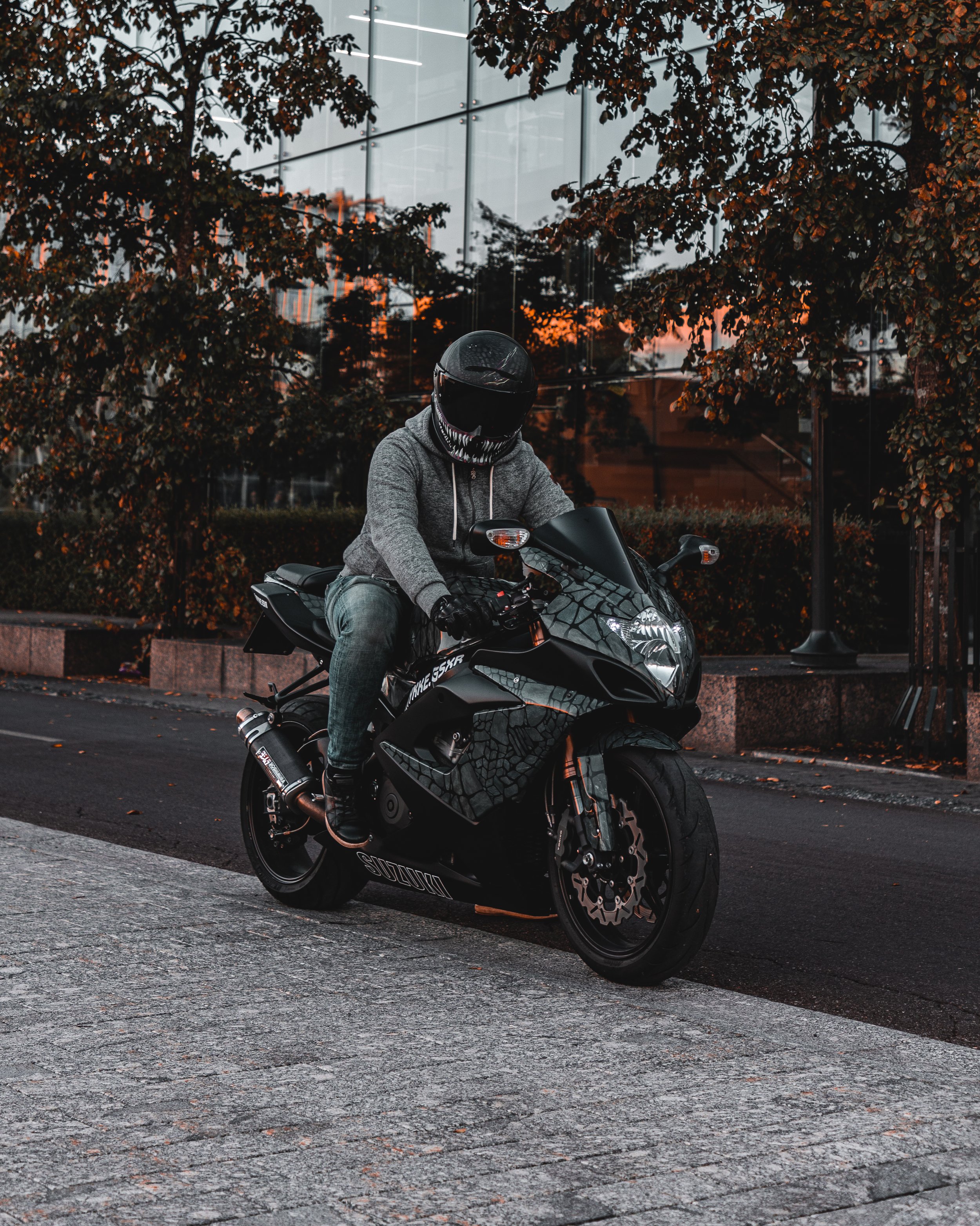 A person riding a black sport motorcycle on a city street during sunset. The rider is wearing a gray hoodie, jeans, black gloves, and a black full-face helmet with a design resembling a skull.