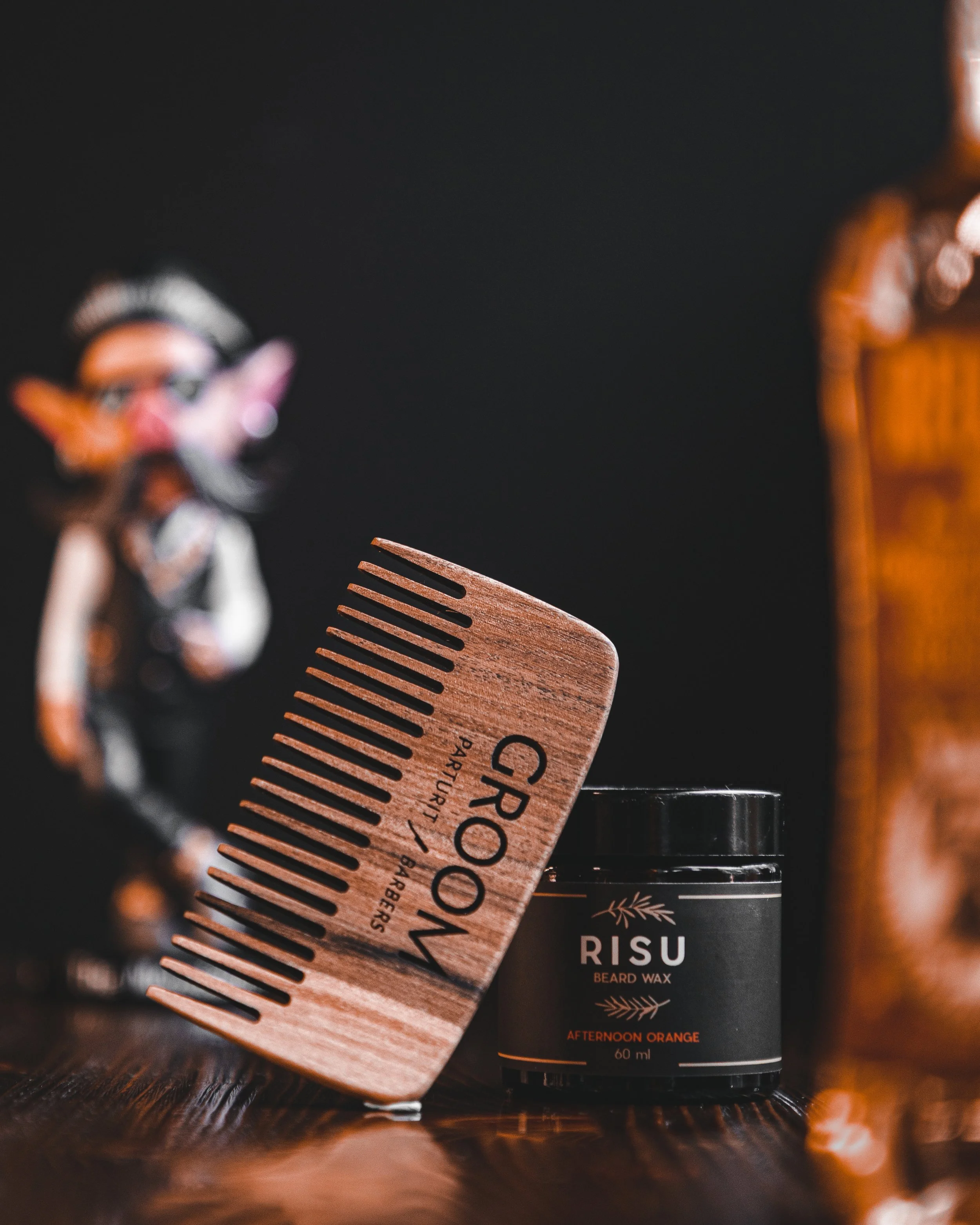 A grooming comb labeled 'Groom' with a barcode design leaning against a jar of RISU beard wax labeled 'Afternoon Orange 60 ml' on a wooden surface, with a blurred figurine in the background.
