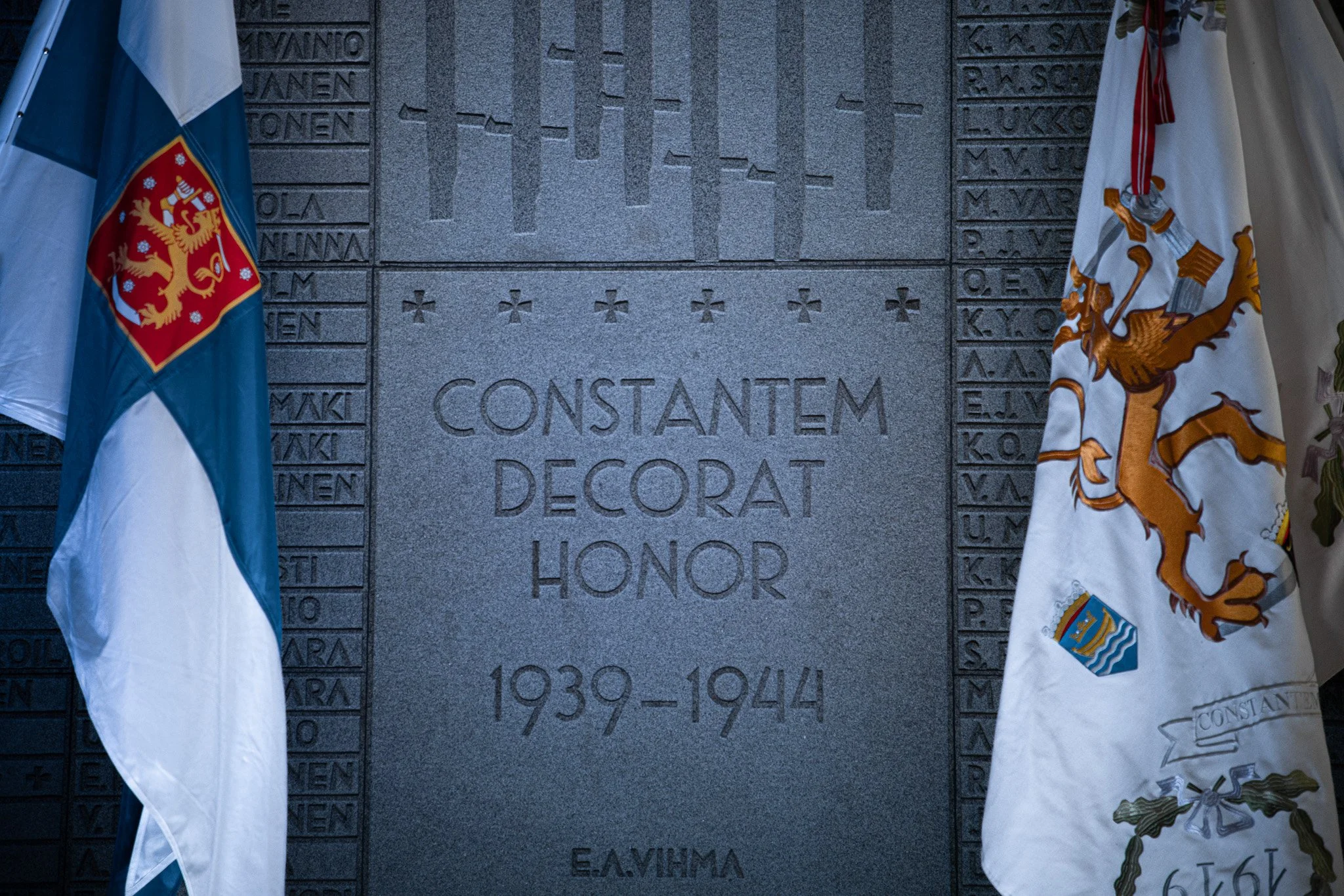 A memorial plaque with the inscription "CONSTANTEM DECORAT HONOR 1939-1944" flanked by two flags, one with a lion crest and the other with a coat of arms, at a dark stone wall with engraved names and a map outline.