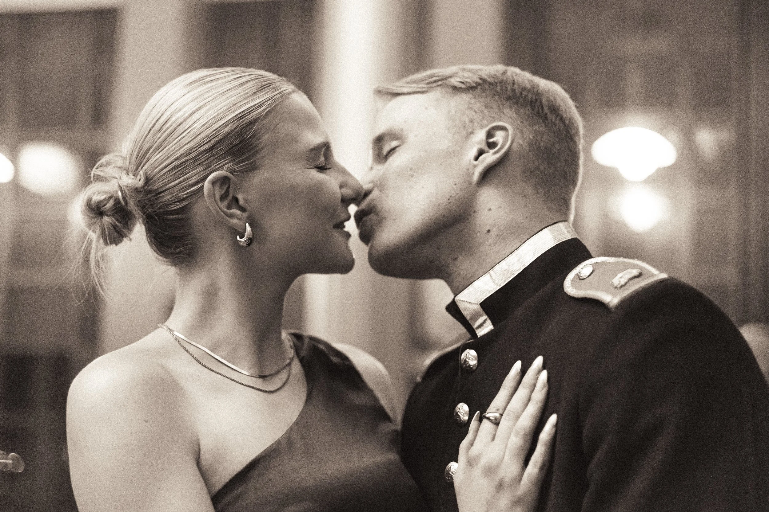A black-and-white photo of a couple about to kiss, with the woman wearing earrings and necklaces, and the man in a military uniform, indoors with blurred lights in the background.