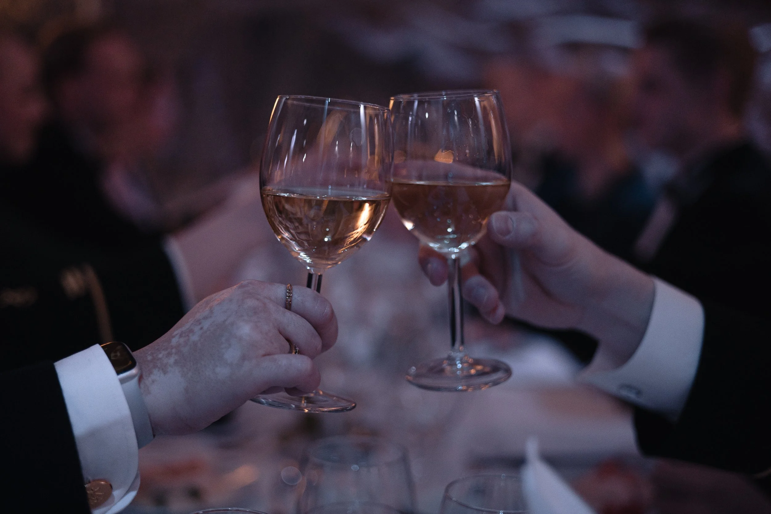 Two people in formal attire clinking glasses of white wine at a celebration or dinner event.