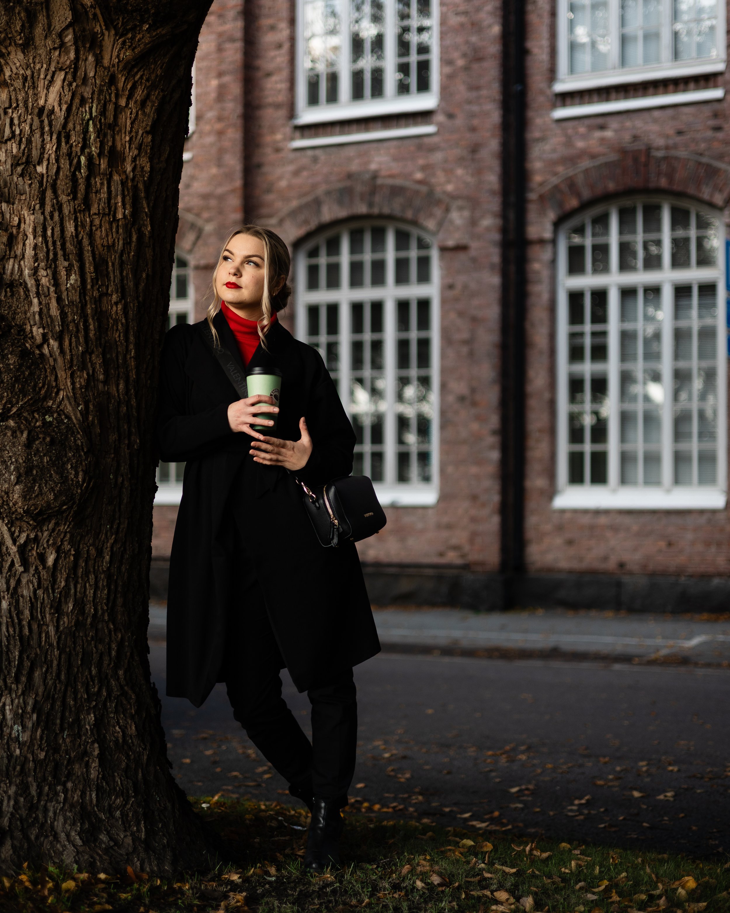 A woman standing under a tree on a city street at dusk, holding a coffee cup, with a brick building and large windows in the background.