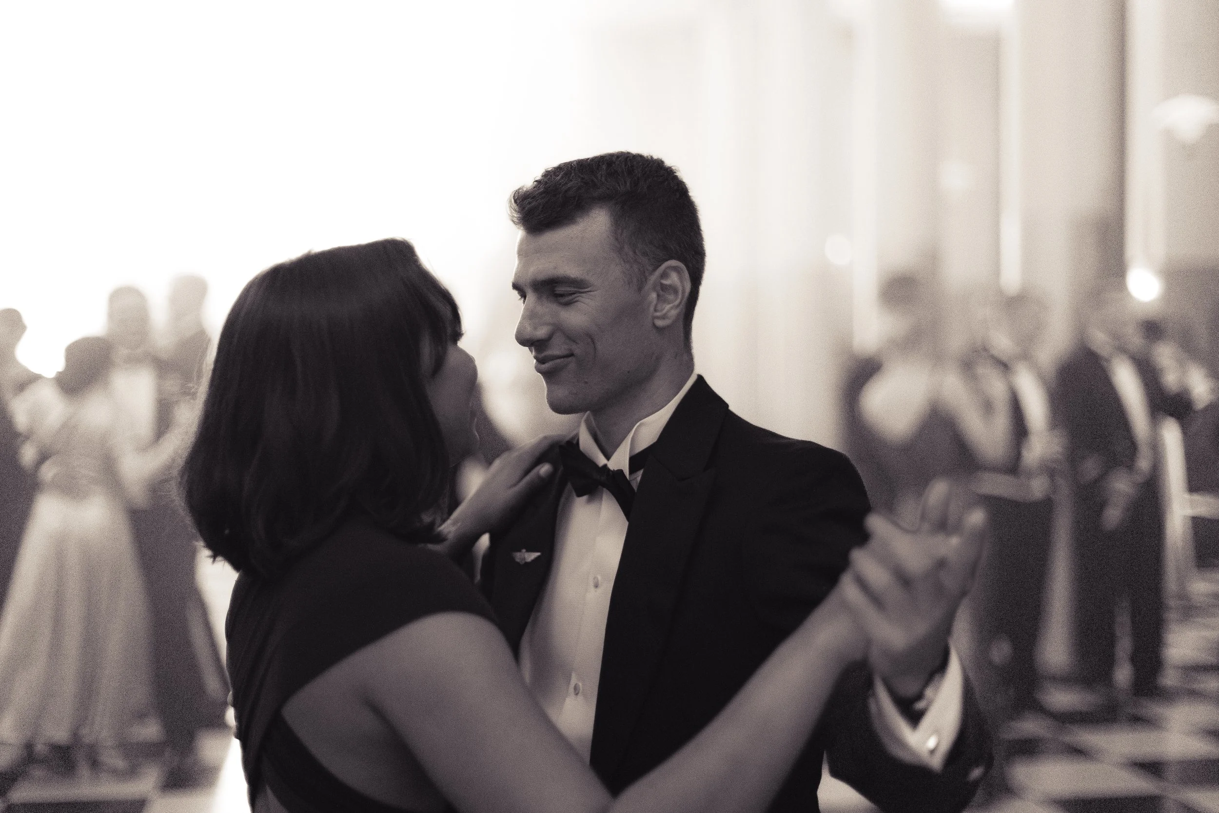 A man in a tuxedo and woman in a dress dancing closely at a formal event with other couples in the background.