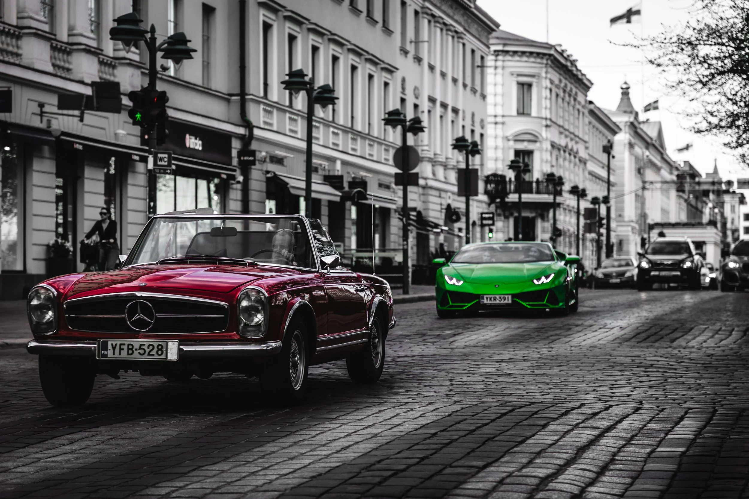 A street scene with a classic red Mercedes-Benz car and a modern green Lamborghini sports car, with black and white surroundings and a few pedestrians.