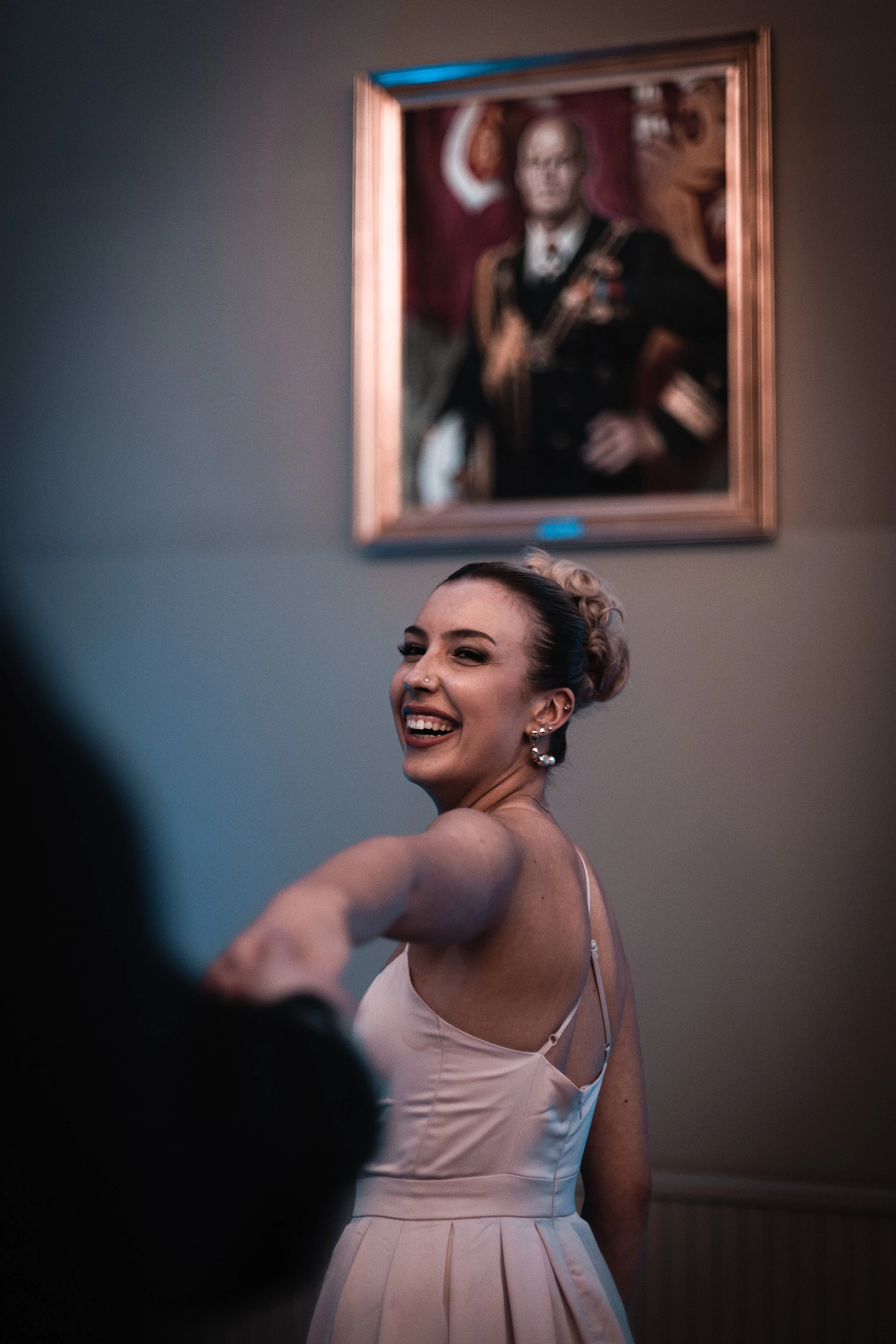A woman with styled hair, earrings, and a light pink dress smiling and reaching out, possibly being spun around, in an indoor setting with a portrait of a man in a military uniform hanging on the wall behind her.