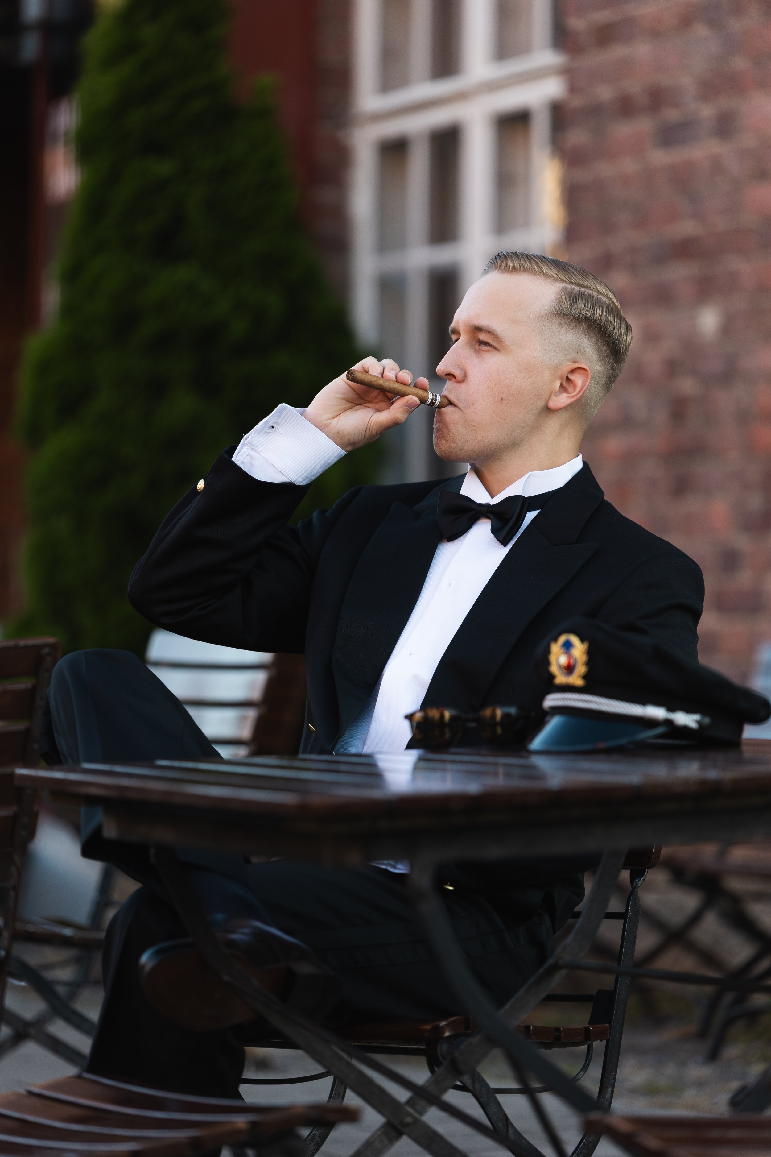 A man in a formal tuxedo with a bow tie, sitting outdoors at a wooden table, smoking a cigar, with a building and greenery in the background.