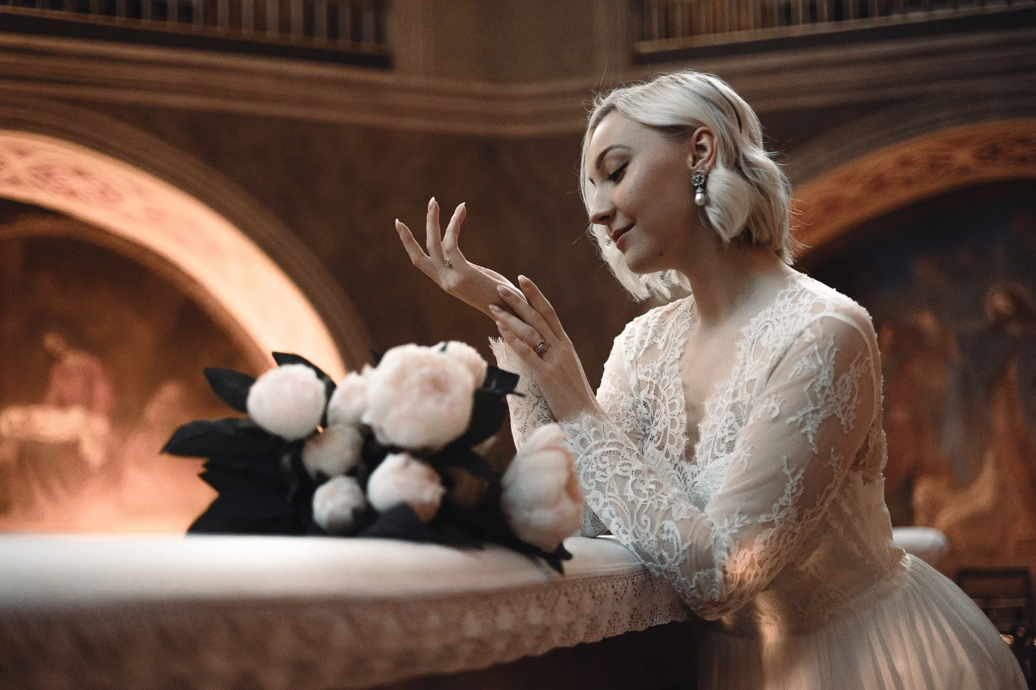 A woman in a lace wedding dress with blonde hair and earrings looks at a bouquet of white flowers on a table in a church or cathedral.