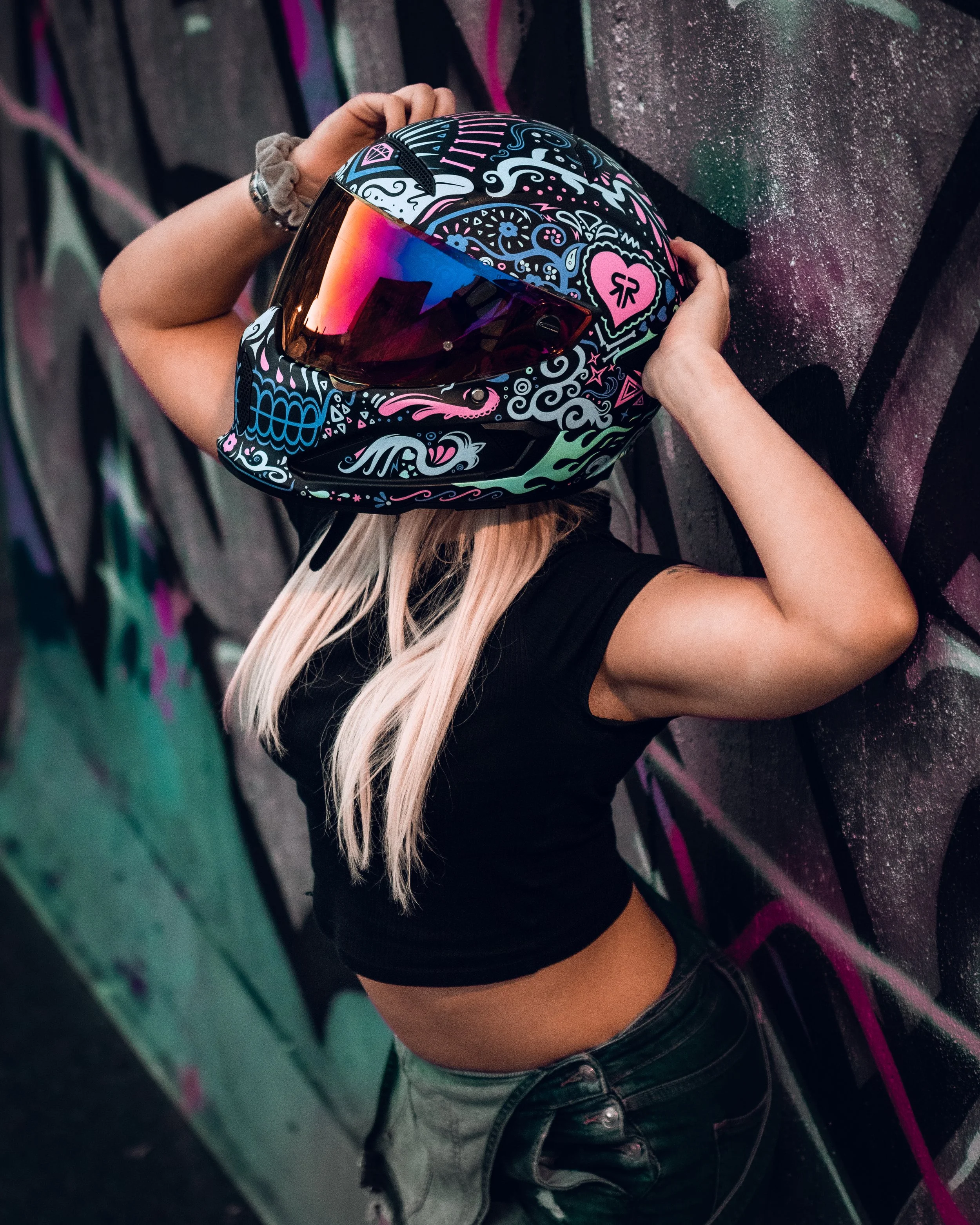 A woman with long blonde hair leaning against a graffiti-covered wall, wearing a black crop top, gray jeans, and a colorful helmet with various patterns and a reflective visor.