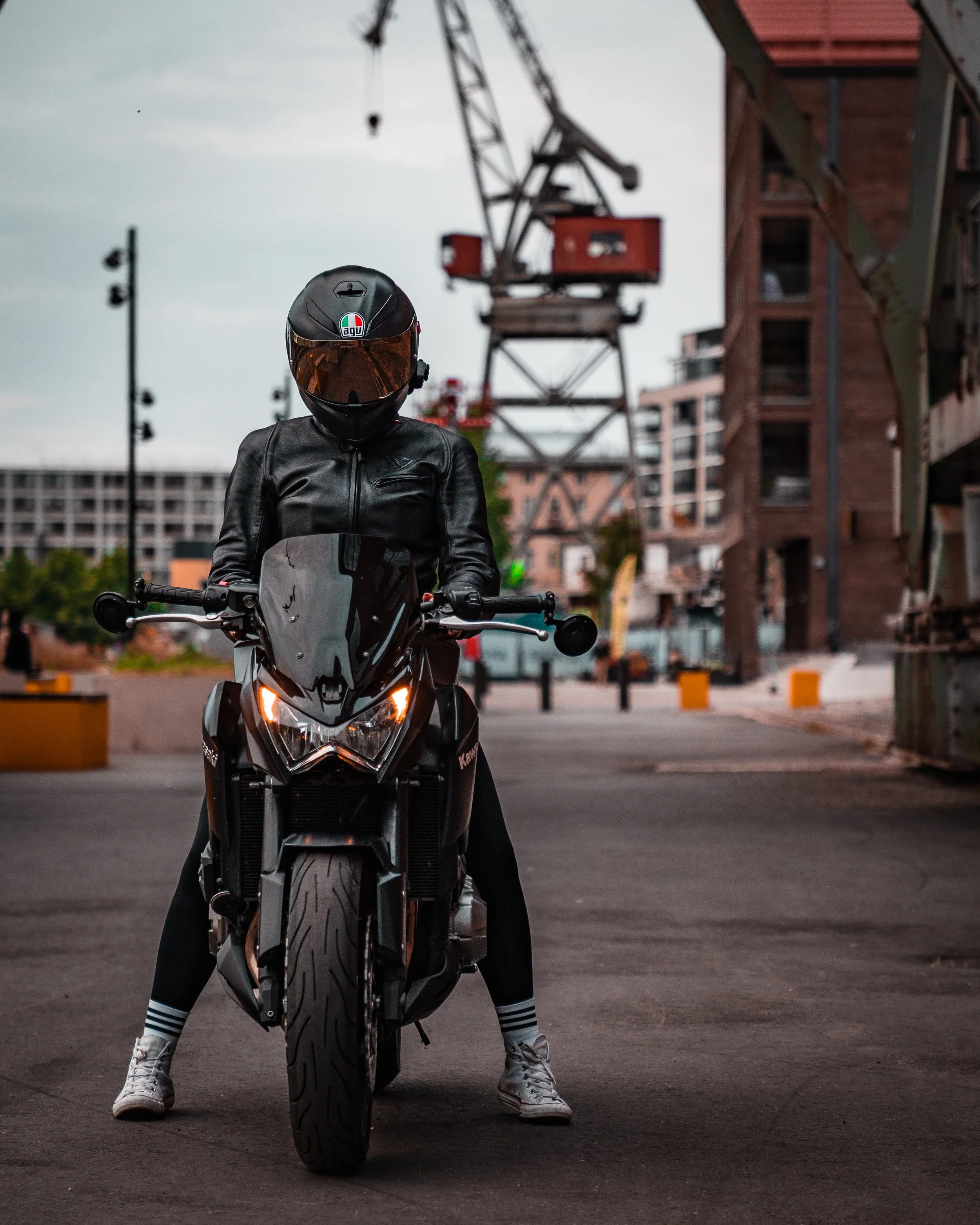Person in black leather jacket and helmet sitting on a black motorcycle on a city street with construction cranes and buildings in the background.