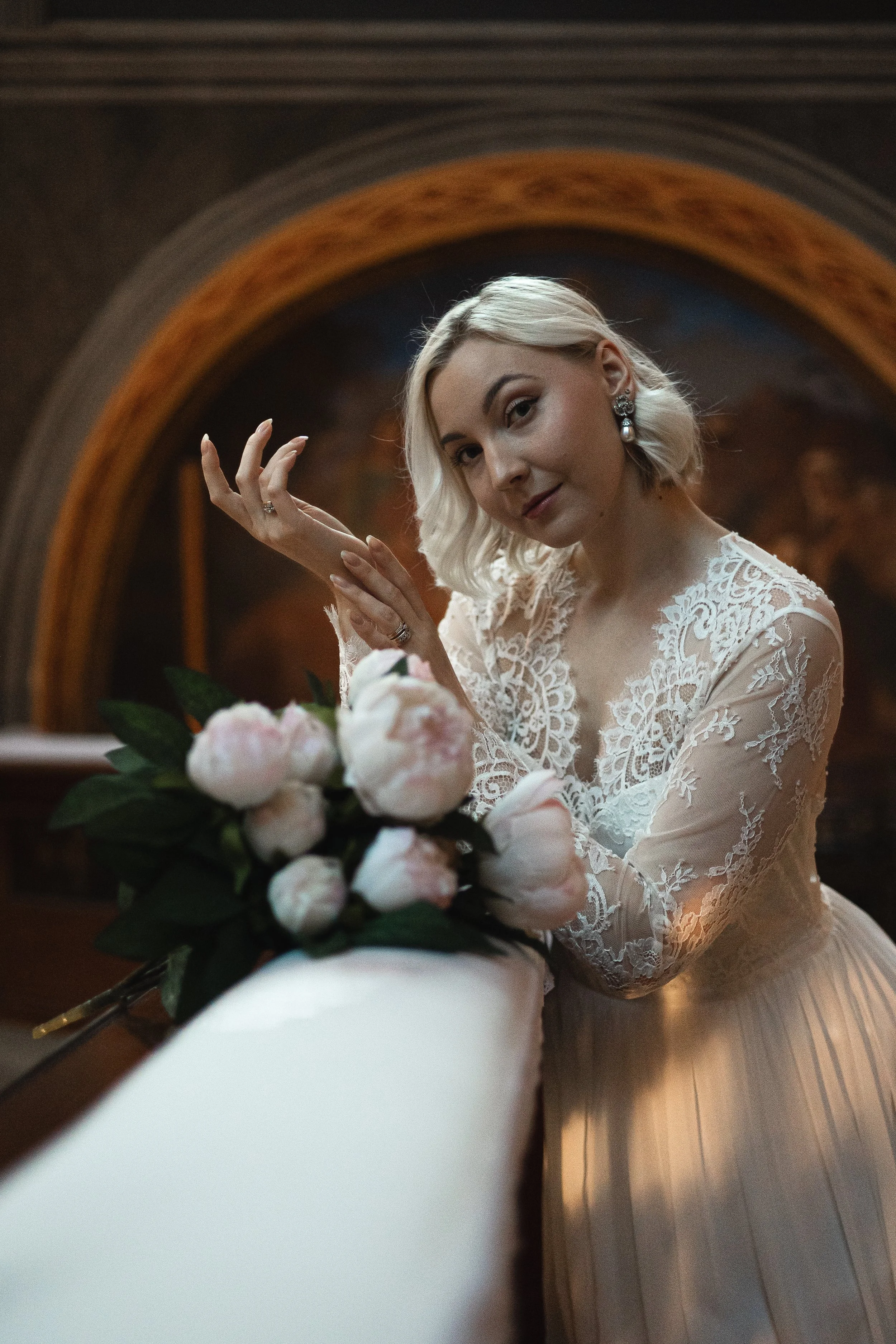 A woman in a white lace wedding dress holding a bouquet of pink peonies, standing in a decorated indoor setting.
