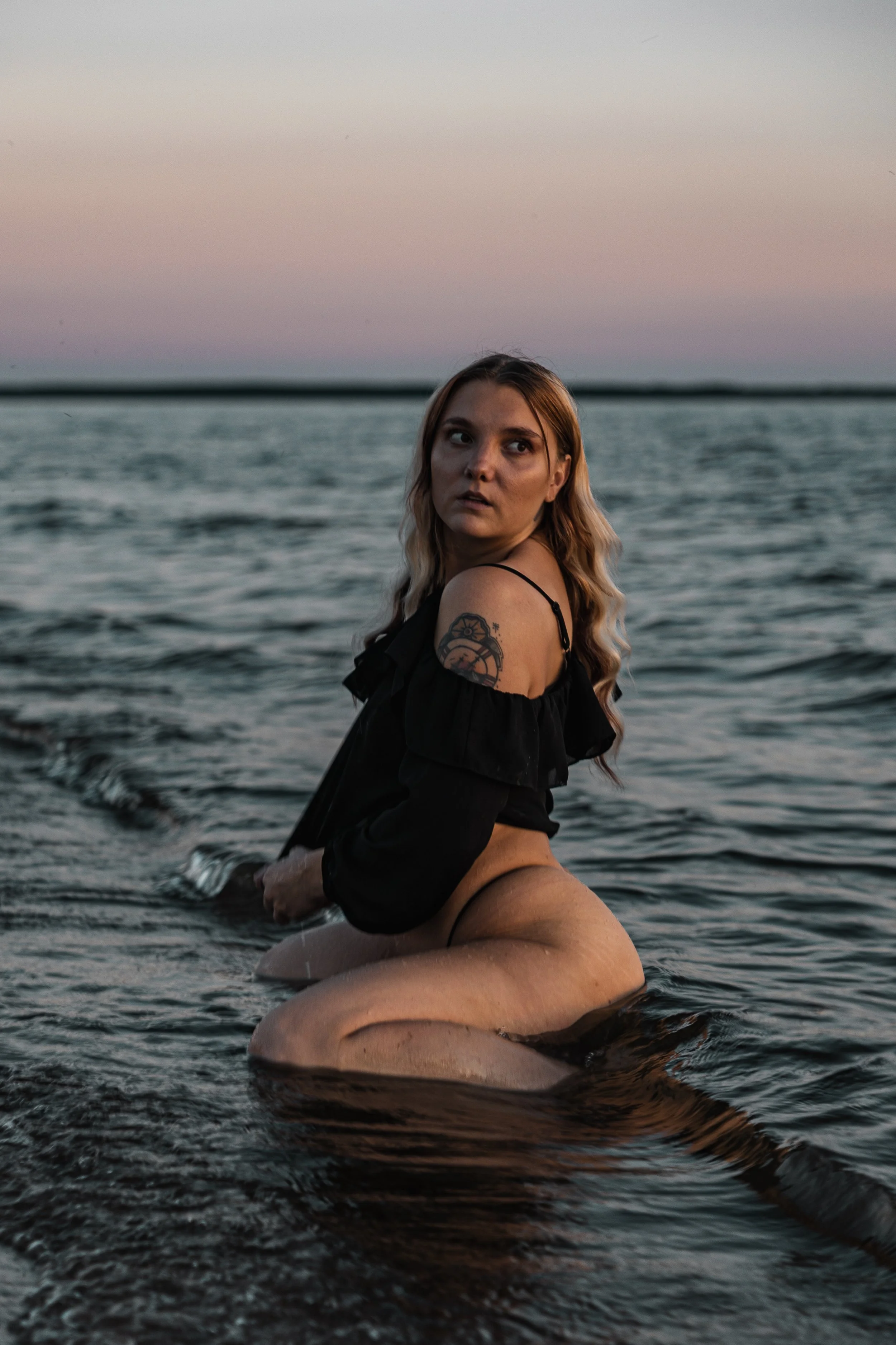 A woman kneeling in the water at sunset, looking over her shoulder with a contemplative expression, wearing a black off-the-shoulder top and underwear, with a tattoo on her upper arm.