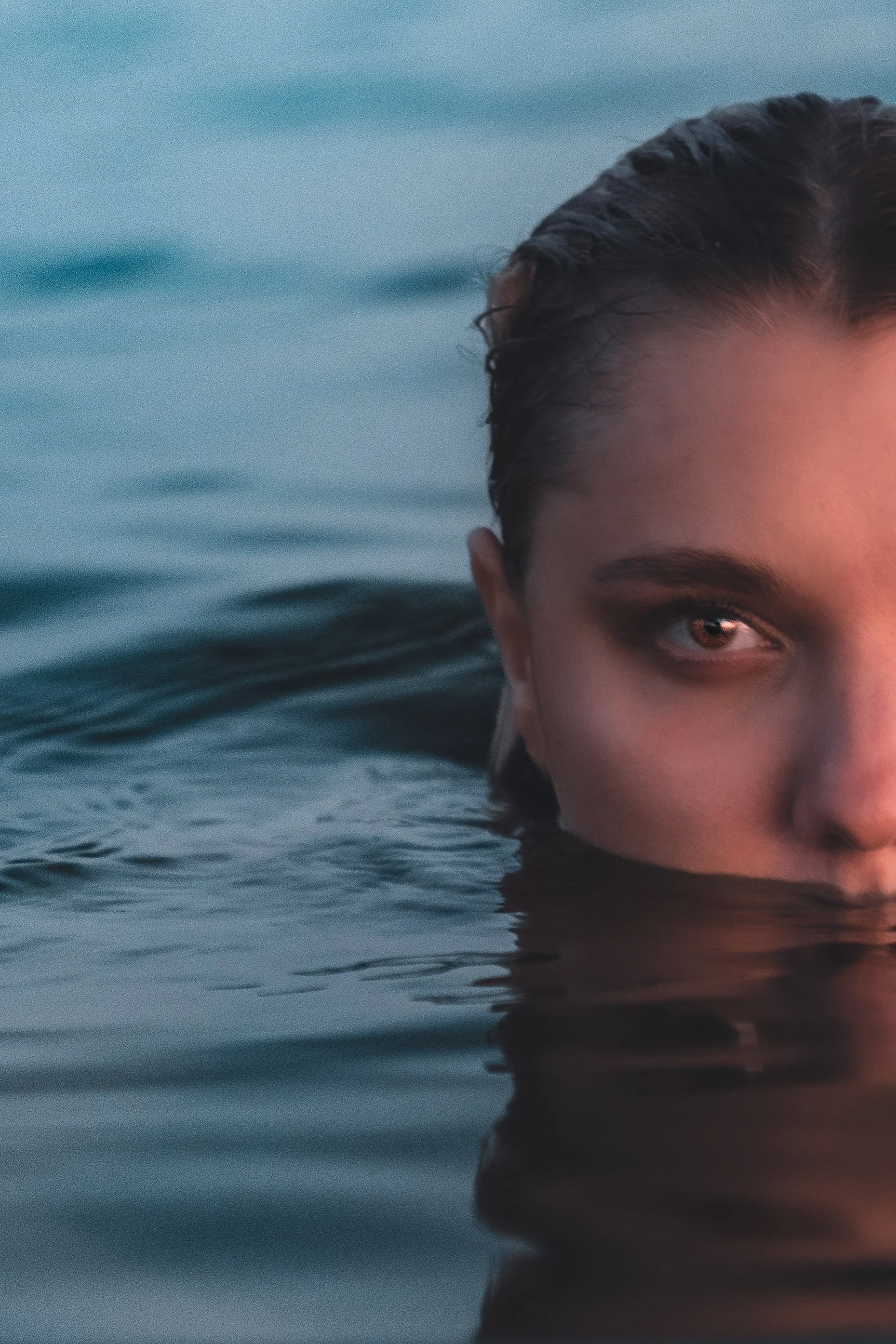 Close-up of a woman's face half submerged in water, with only her right eye, eyebrow, and part of her forehead visible, water reflecting on her skin.